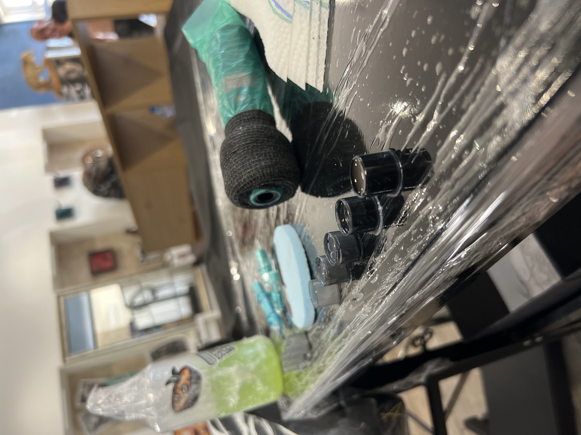 The image shows a close-up of tattoo equipment set on a black surface covered in plastic wrap. A tattoo machine, wrapped in green protective plastic and with a textured black grip, is present. Next to it are several small, black ink caps. Other items visible include a blue sponge and a bottle with a green liquid, likely cleaning or sterilizing solution. The scene appears to be a tattoo studio setup.
In the blurred background, there is a wooden shelving unit with various items stored within. There is a person in the upper left, presumably a tattoo artist. The environment appears to be a professional space. There are no clear indicators that definitively confirm the location as Sofia, Bulgaria.