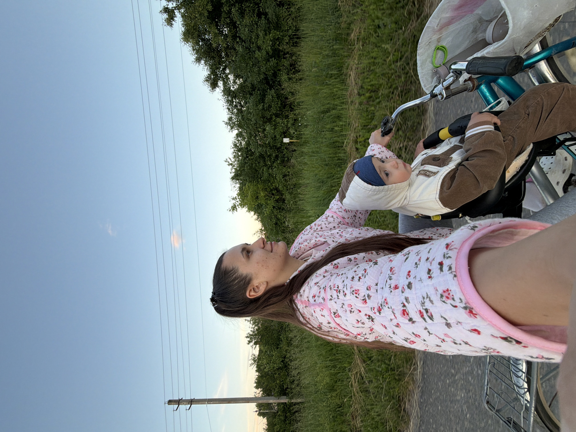 A woman and a child are riding on a bicycle. The woman, with her hair in a ponytail, is wearing a pink floral-patterned robe. She is looking to her right, with her arm extended forward as if taking a selfie. The child, wearing a brown and white hooded jacket and a blue cap, is seated in a child seat attached to the front of the bicycle. The child is looking forward. They are on a paved road, with a grassy field and trees in the background. A utility pole with overhead wires is visible on the left side of the image. The sky is clear blue with a hint of a cloud. The scene appears to be set in a rural area, possibly on the outskirts of Красный Маяк, Russia, given the context provided. The lighting suggests it is either early morning or late afternoon.