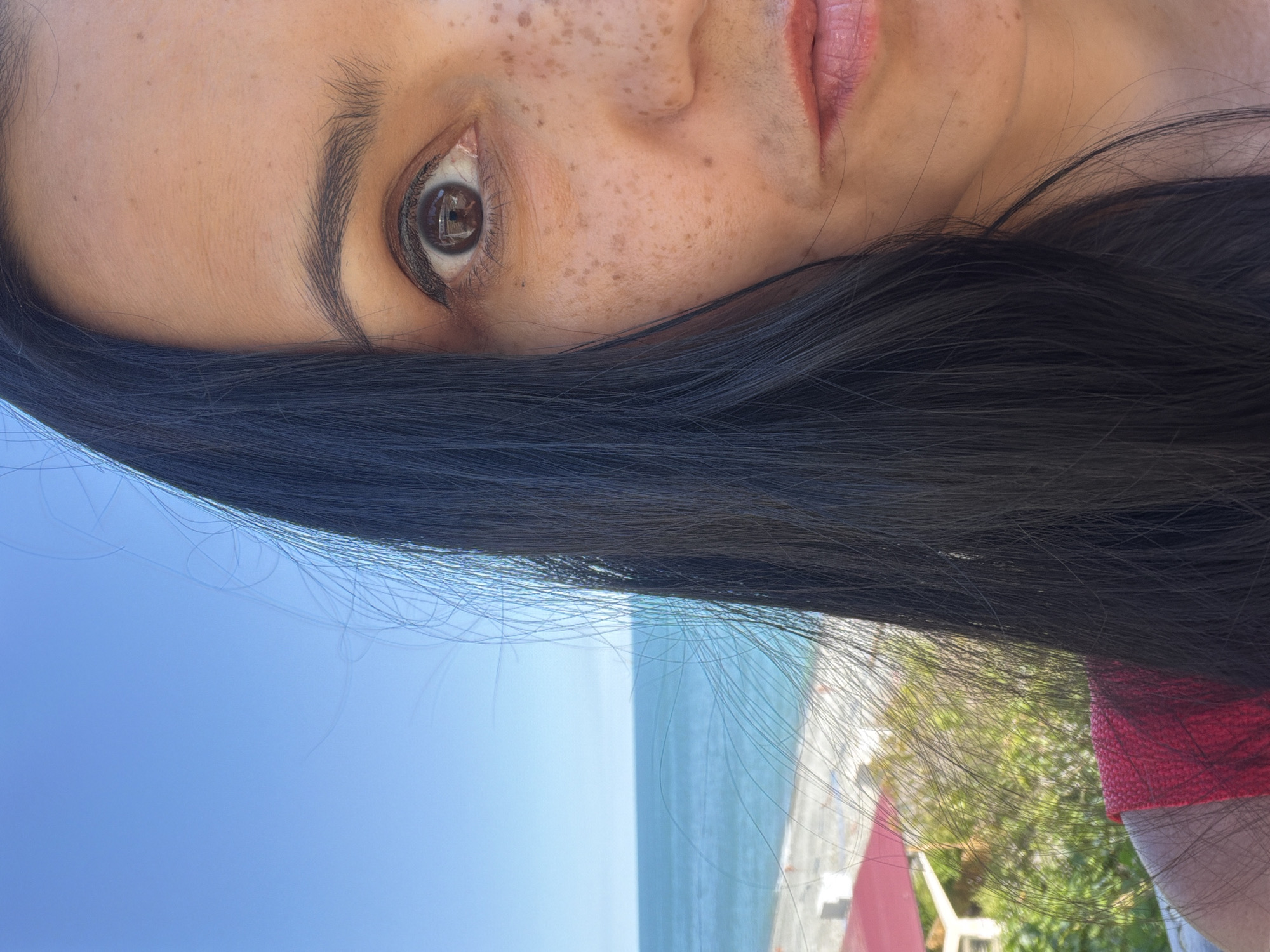 The media file is a close-up, vertically oriented selfie of a person's face, showing their eye, nose, mouth, and hair. The subject has fair skin with freckles across their nose and forehead. They have dark, long hair that partially obscures their face. Their eye is brown and appears to be looking directly at the camera, with visible eyelashes and eyebrows. The mouth is slightly open, revealing pink lips.
In the background, a bright blue sky transitions into a turquoise ocean with a sandy beach and some greenery visible. There are also hints of what appears to be beach furniture or structures in the distance, suggesting an outdoor setting, likely a beach or seaside resort. The lighting indicates it is daytime, likely a sunny day. The overall impression is one of relaxation and enjoyment of a pleasant outdoor environment. The location is specified as Sochi, Russia. No visible text is present in the media file.