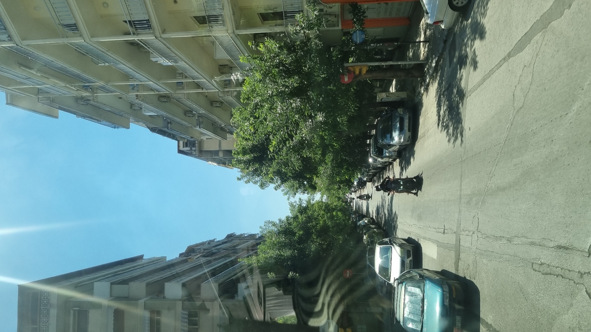 The image shows a street in the city of Thessaloniki Municipal Unit, Greece, on a sunny day. The street is lined with apartment buildings on both sides, with balconies and air conditioning units visible. Dense green trees cast shadows on the road and sidewalk. Several cars are parked along the sides of the street. A man on a scooter is riding down the middle of the road, with another scooter slightly ahead of him. The sky is clear blue with bright sunlight streaming down, creating lens flare effects.