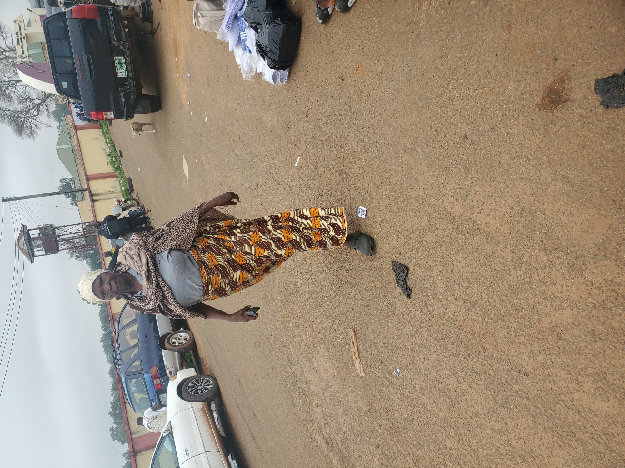 The image shows an outdoor scene with an elderly woman in the foreground, facing the camera. She is wearing a headscarf, a patterned scarf draped over her shoulders, a grey t-shirt, and a long, patterned skirt. Her right hand is raised, holding a dark object, possibly a phone or wallet. Her left hand is by her side. She is wearing dark shoes.
Behind the woman, there are several vehicles including a white car on the left and a blue minivan. Further in the background, a black pickup truck is parked. A small dog is visible near the truck. Beyond the vehicles, there is a building with a fenced-in area and a guard tower. The ground is paved with asphalt, and there are various items scattered on it, including black plastic bags and what appears to be white fabric or clothing.
The environment suggests a public area, possibly a marketplace or a street near a facility. The sky is overcast, indicating a cloudy day. The overall scene is of everyday life in what appears to be a somewhat busy, outdoor location. The location is identified as Bahit, Nigeria.