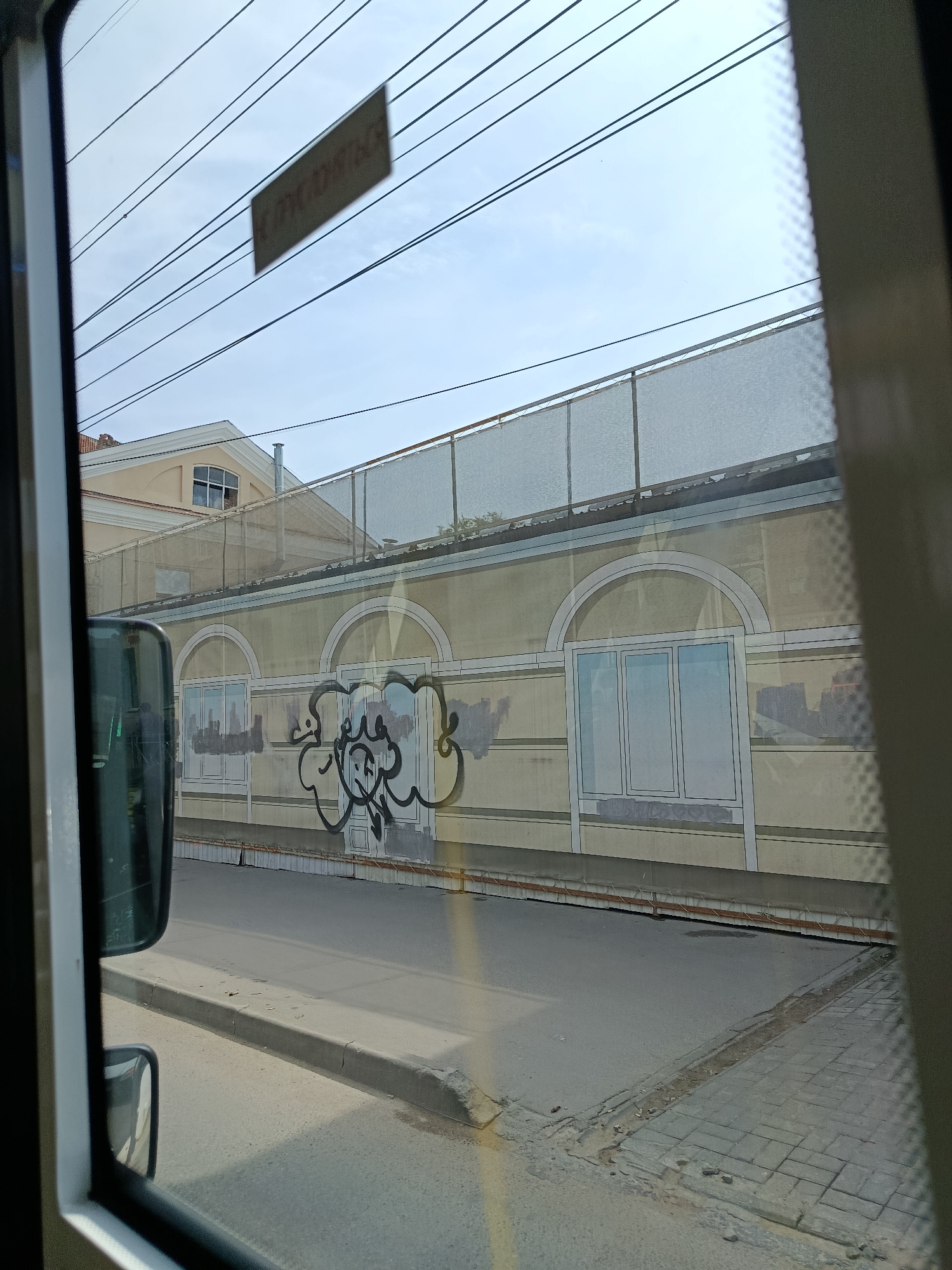 The image, taken from inside a vehicle, captures a view of a building exterior and a street. The building facade is painted in a pale yellow color and features arched window frames, some of which are depicted as windows and others as painted trompe-l'oeil elements. Graffiti in black spray paint is visible on the lower section of the building, obscuring some of the painted architectural details. A mesh-like screen is stretched across the upper part of the building, possibly for construction or protection. Overhead, power lines crisscross the sky. A sign, seemingly rectangular and dark, is affixed to the wires. The street in front of the building is paved, with a curb and sidewalk. A side-view mirror of the vehicle from which the photo is taken is partially visible on the left. The overall impression is an urban scene, likely in Saratov, Russia, given the context.