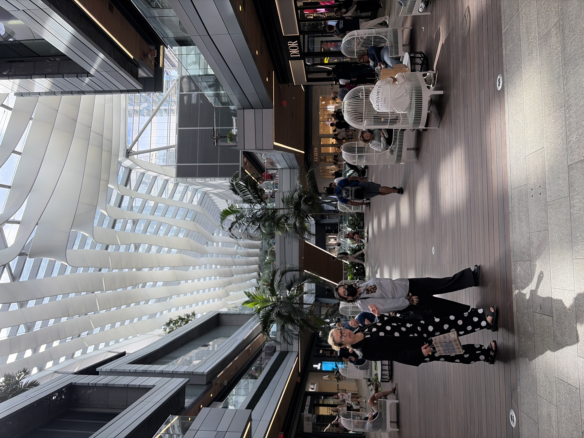 The photograph depicts an interior view of a modern shopping mall, likely located in Miami, United States, considering the presence of palm trees and architectural style. The roof is a distinctive, undulating white structure, providing natural light. Below the roof, the mall features multiple floors with stores visible on both sides. Several shoppers are present, some walking along the wooden-planked walkway. Storefronts such as "DIOR" are visible. People are seated in white, cage-like chairs. Two individuals, one wearing a black polka-dot outfit and the other in a grey sweatshirt and black pants, are walking toward the viewer.