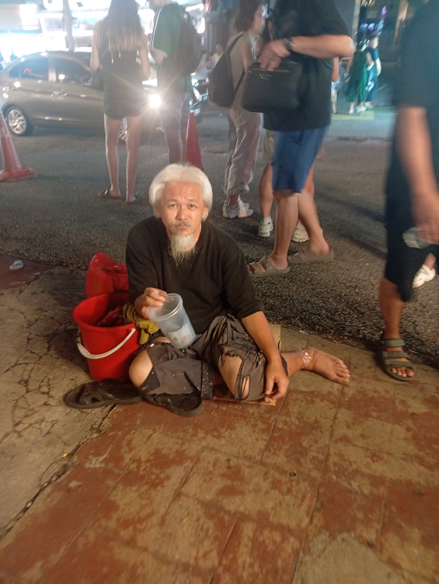 The image depicts a scene on a street at night, likely in Kuala Lumpur, Malaysia, given the context of the surrounding urban environment with illuminated buildings and traffic. In the foreground, a man with gray hair and a goatee is sitting on the ground, holding a clear plastic cup. He is wearing a dark long-sleeved shirt and ripped, dark-colored shorts. Next to him is a red bucket, and to his left are a pair of black flip-flops. His left leg is bent, and his right leg is extended. The ground appears to be a mix of pavement and wooden planks. In the background, several people are walking on the street. A silver car is visible on the left side of the image, and there are traffic cones. The lighting suggests an urban setting with artificial illumination from streetlights and buildings.