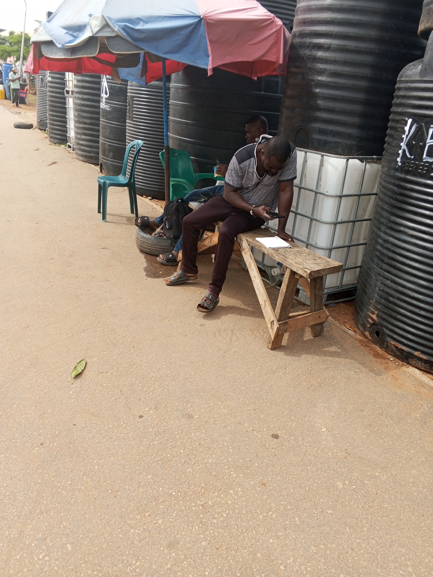 The image shows a street scene in Abuja, Nigeria. A man sits on a wooden bench, looking at a cell phone. A notebook rests on the bench next to him. Another man sits nearby on a tire, leaning against a large white container placed amongst several large, black water tanks. A large multi-colored umbrella provides shade. Several plastic chairs are nearby. A single leaf lies on the ground.