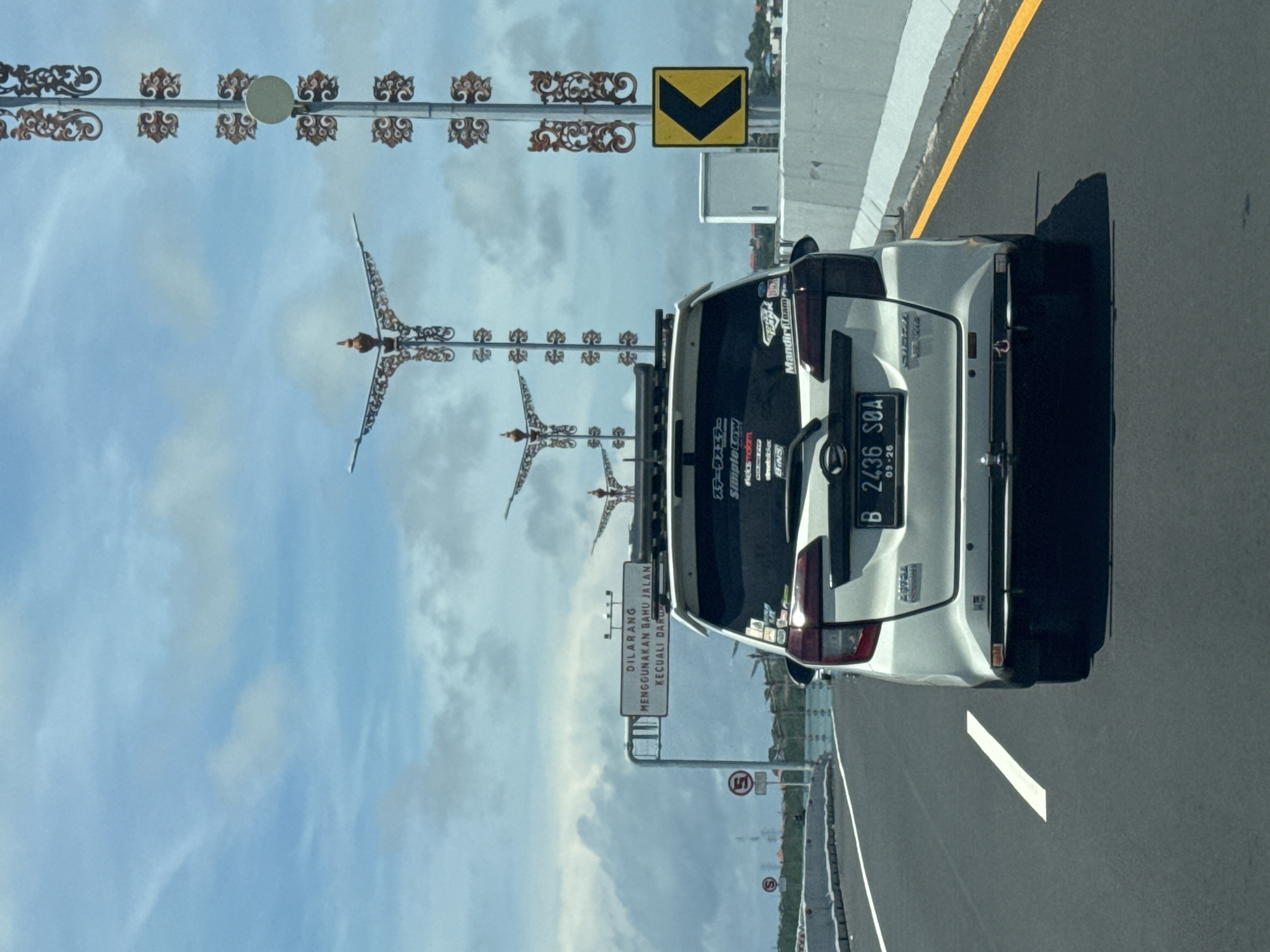 A white van is on a highway near Nusa Dua, Indonesia. The vehicle is in the right lane, with a white road line to the left. The van has a roof rack, stickers on the back window, and a license plate that reads "B 2436 SOA". A road sign to the left of the van warns, "DILARANG MENGGUNAKAN BAHU JALAN KECUALI DARU," meaning "DO NOT USE THE SHOULDER OF THE ROAD EXCEPT IN EMERGENCIES." Decorative light poles with bird-shaped elements line the highway's edge, and a yellow chevron sign is visible near the concrete barrier. The sky is overcast with clouds.