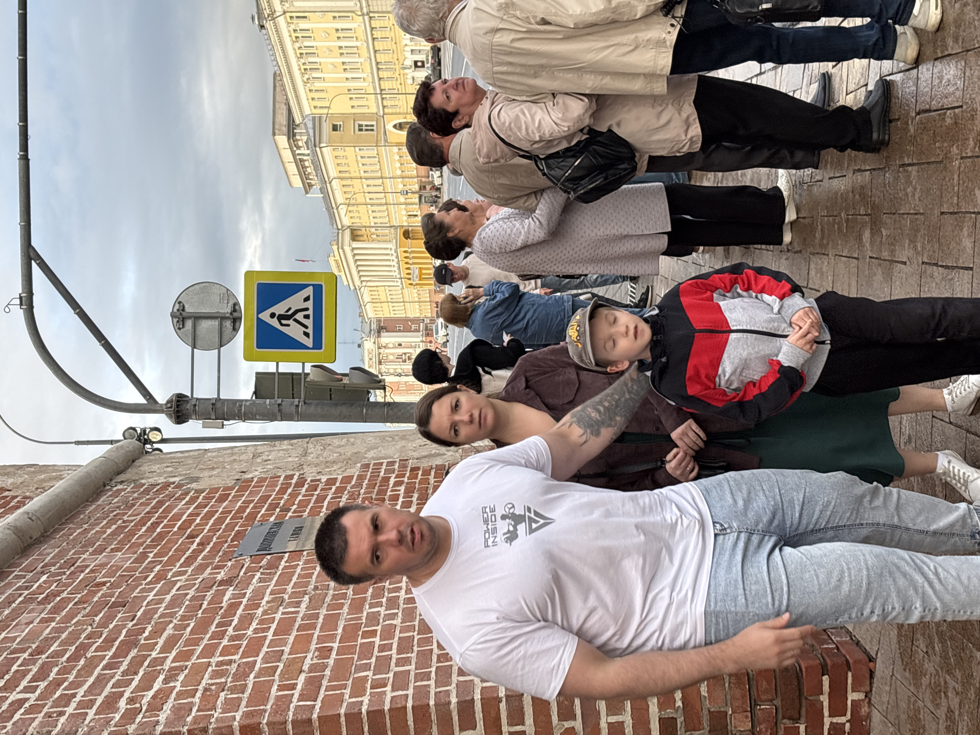 On a cloudy day in Nizhny Novgorod, Russia, a group of people stand on a sidewalk next to a brick building and a street. The man in the foreground, wearing a white t-shirt and light blue jeans, has a tattoo on his arm. A young boy in a colorful jacket and a woman in a dark green dress are also visible. Other pedestrians, dressed in casual attire, are waiting behind them. A pedestrian crossing sign is displayed on a pole. In the background, a yellow building with classical architecture lines a wide street. The ground is wet, suggesting recent rain.
