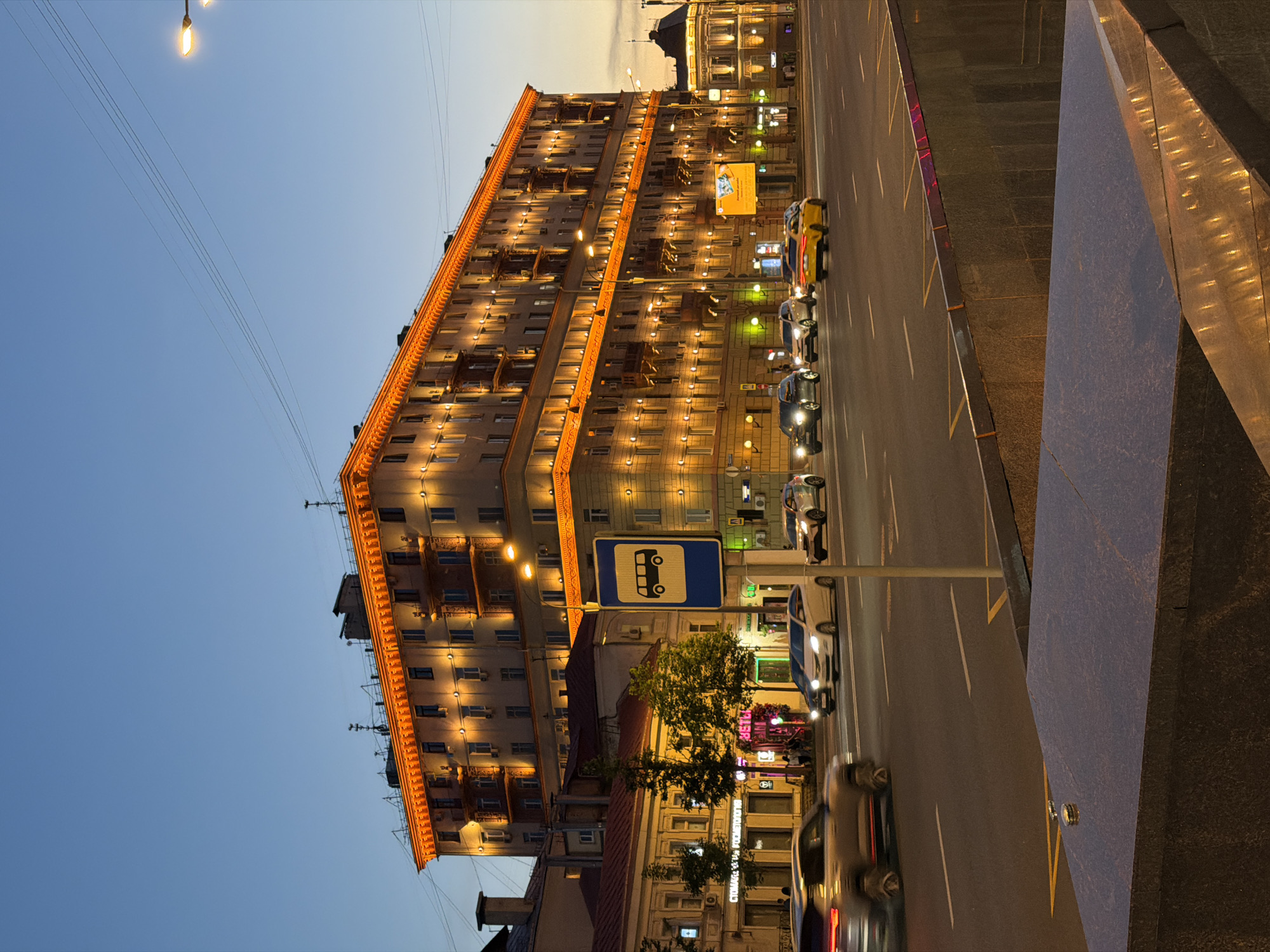 This image captures an urban street scene in Moscow, Russia, at dusk or early evening. The sky transitions from a deep, clear blue at the top to a lighter, amber hue near the horizon on the right, indicating a recent sunset.
The dominant feature is a large, multi-story building, likely an apartment or mixed-use complex, occupying the left and central background. This cream-colored structure, characteristic of Stalinist Empire architecture, is extensively illuminated with warm orange lights along its cornices, window ledges, and between floors, highlighting its architectural details, including numerous windows and balconies. Antennas and other structures are visible on its rooftop.
In the mid-ground, a wide asphalt road with multiple lanes traverses the scene. Several vehicles are present: a silver SUV is blurred in motion in the immediate foreground on the left, while further back, a white sedan, another silver SUV, a yellow taxi, and a dark car are visible, all with their headlights on. White lane markings and yellow diagonal lines on the road surface are discernible.
On the left side of the road, a blue square sign with a white bus icon indicates a bus stop, mounted on a pole. Behind this sign, smaller, older-style buildings with red roofs are partially visible, featuring illuminated storefronts. One sign on a building reads "СТОМАТОЛОГИЯ" (Dentistry), and another appears to say "ЦВЕТЫ" (Flowers). A small group of people can be seen near one of these brightly lit storefronts. On the far right, another building with warm architectural lighting is partially visible in the distance. Overhead utility wires and streetlights with warm glows are also part of the urban infrastructure.
The immediate foreground features a dark, polished stone structure, likely part of an underpass entrance or a street barrier, with some reflective surfaces catching the ambient light.
The scene depicts a bustling urban environment illuminated for the evening, specifically identifiable as **Tverskaya Street (Тверская улица)** in Moscow, Russia, given the distinctive architecture, wide avenue, and general urban layout.