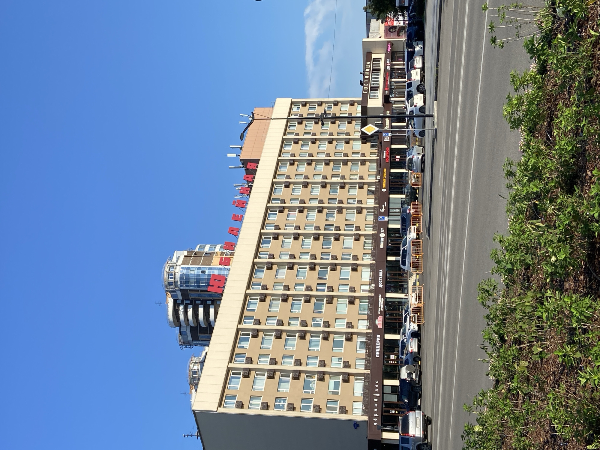 The image shows a large, tan-colored hotel building with numerous windows and air conditioning units visible on its facade. Red Cyrillic lettering spelling "ЮБИЛЕЙНАЯ" (Yubileynaya) is prominently displayed across the top of the hotel, indicating it is likely the "Yubileynaya" hotel. A modern, multi-story building with rounded balconies and glass features stands behind and to the left of the hotel. The ground floor of the hotel features a row of shops with signs in Cyrillic, including "ЧЁРНЫЙ ЛЕС" (Cherny Les), "ПИЦЦЕРИЯ" (Pizzeria), and "ДОСТАВКА" (Delivery). Cars are parked in front of these establishments, and a street with multiple lanes and a divided median runs in the foreground. Green foliage is planted in the median strip. A blue sky with a few wispy clouds forms the backdrop. The building and signage suggest the location is within the city of Blagoveshchensk, Russia.
