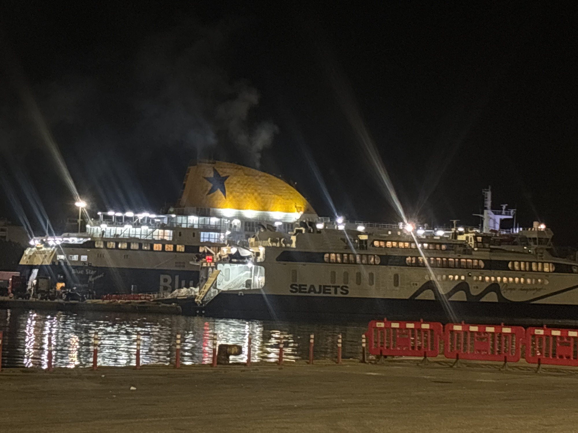 The image shows two large ferries docked at night in what is likely Piraeus, Greece. The ferry on the left is partially visible and has "Blue Star" written on it. The ferry on the right is a "SEAJETS" vessel. Steam or smoke is rising from the top of the ferry. The scene is illuminated by the ship's lights and surrounding streetlights, which reflect on the water. Red and white barriers stand on the dock in the foreground. The surrounding environment is dark, suggesting it's night time.