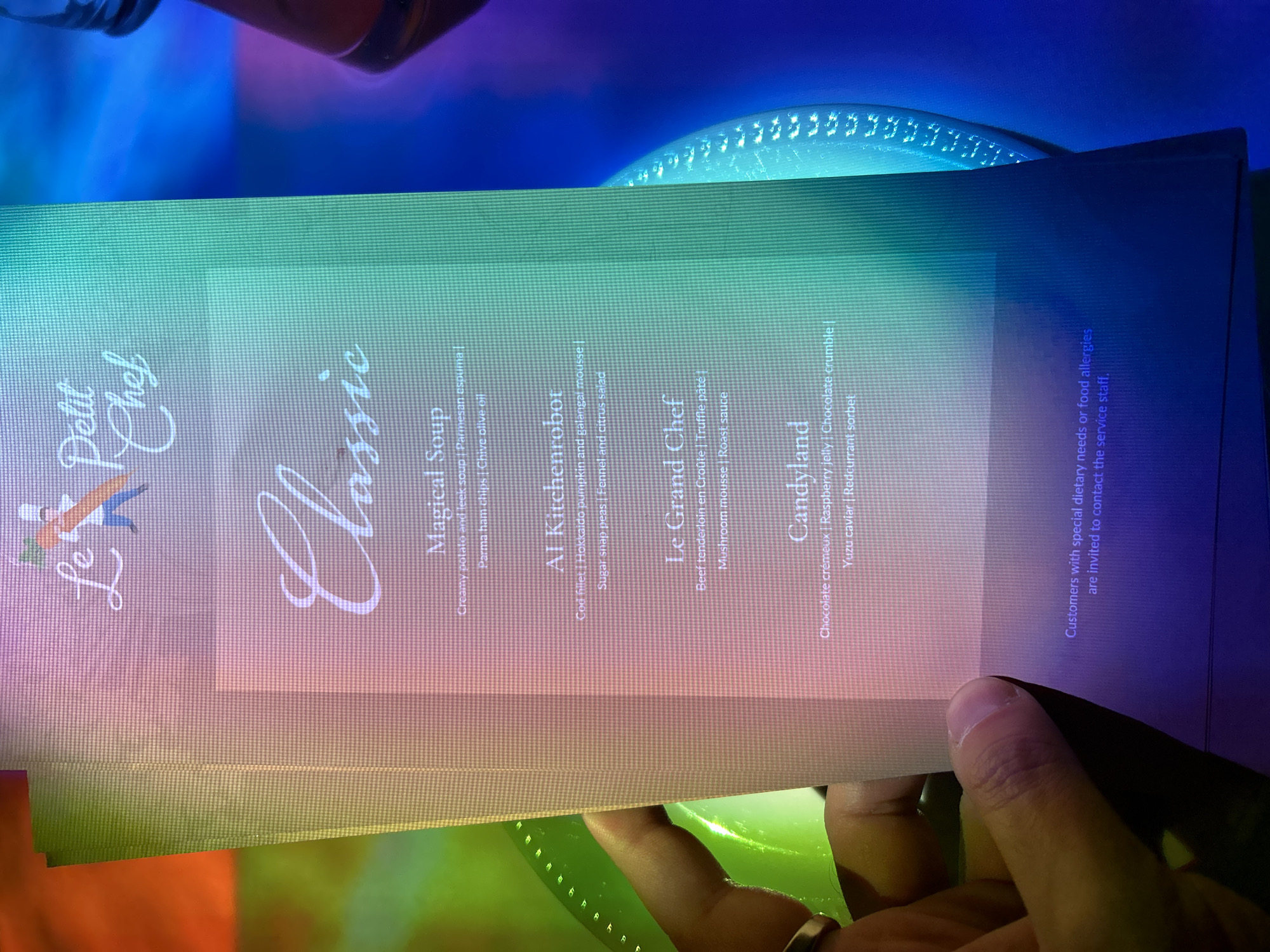 The image shows a Le Petit Chef menu, featuring classic dishes such as Magical Soup, Al Kitchenrobot, Le Grand Chef, and Candyland. It indicates customers with dietary needs should contact the staff. The menu provides detailed descriptions of each dish's components. The location is not revealed in the image, and there is no indication of Warsaw, Poland.