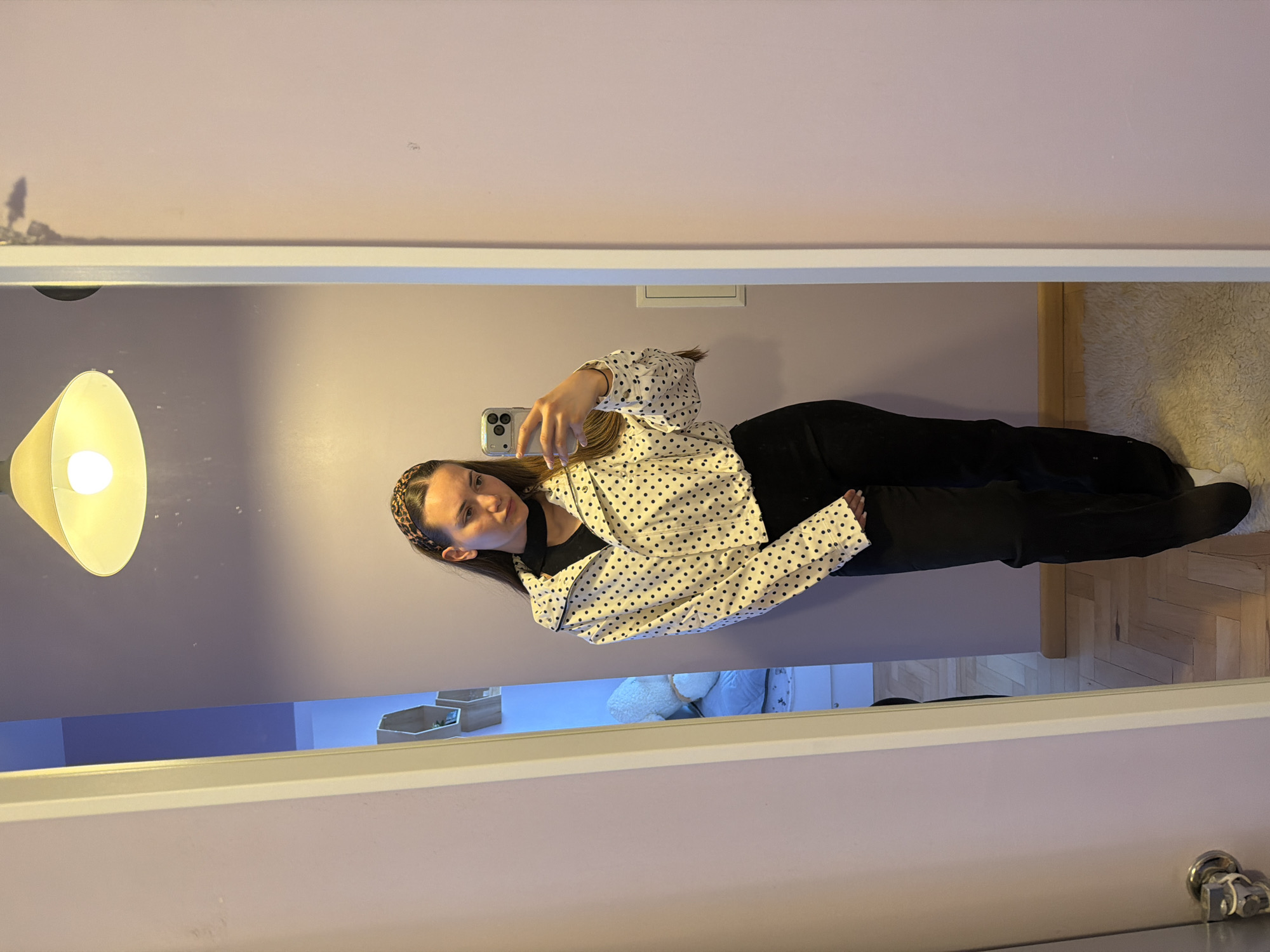 A person is reflected in a mirror, taking a selfie. The individual is wearing a white jacket with black polka dots and black pants. They have long brown hair and a patterned headband. In the background, there is a light fixture, a light purple wall, and a glimpse of a room with items on shelves. The floor appears to be wooden. There is no indication of the location being Buhovo, Bulgaria.