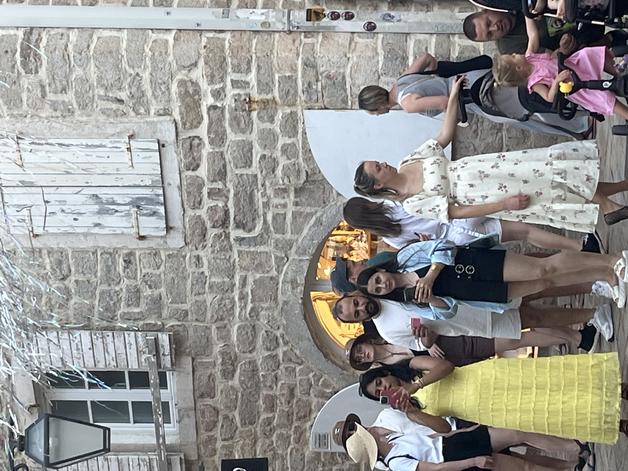 In Budva, Montenegro, a group of people are gathered outside a building with a stone facade. The building has shuttered windows and an arched entrance. A woman in a yellow dress holds a smartphone, and another woman in a light blue shirt and black shorts is also looking at her phone. In the foreground, a young girl in a pink dress is seated in a stroller. To the right, a man wearing a green shirt is visible, and further to the right, another man is seen in the background. The scene appears to be a street or pedestrian area, possibly near shops or restaurants, as suggested by the illuminated interior visible through the arched entrance.