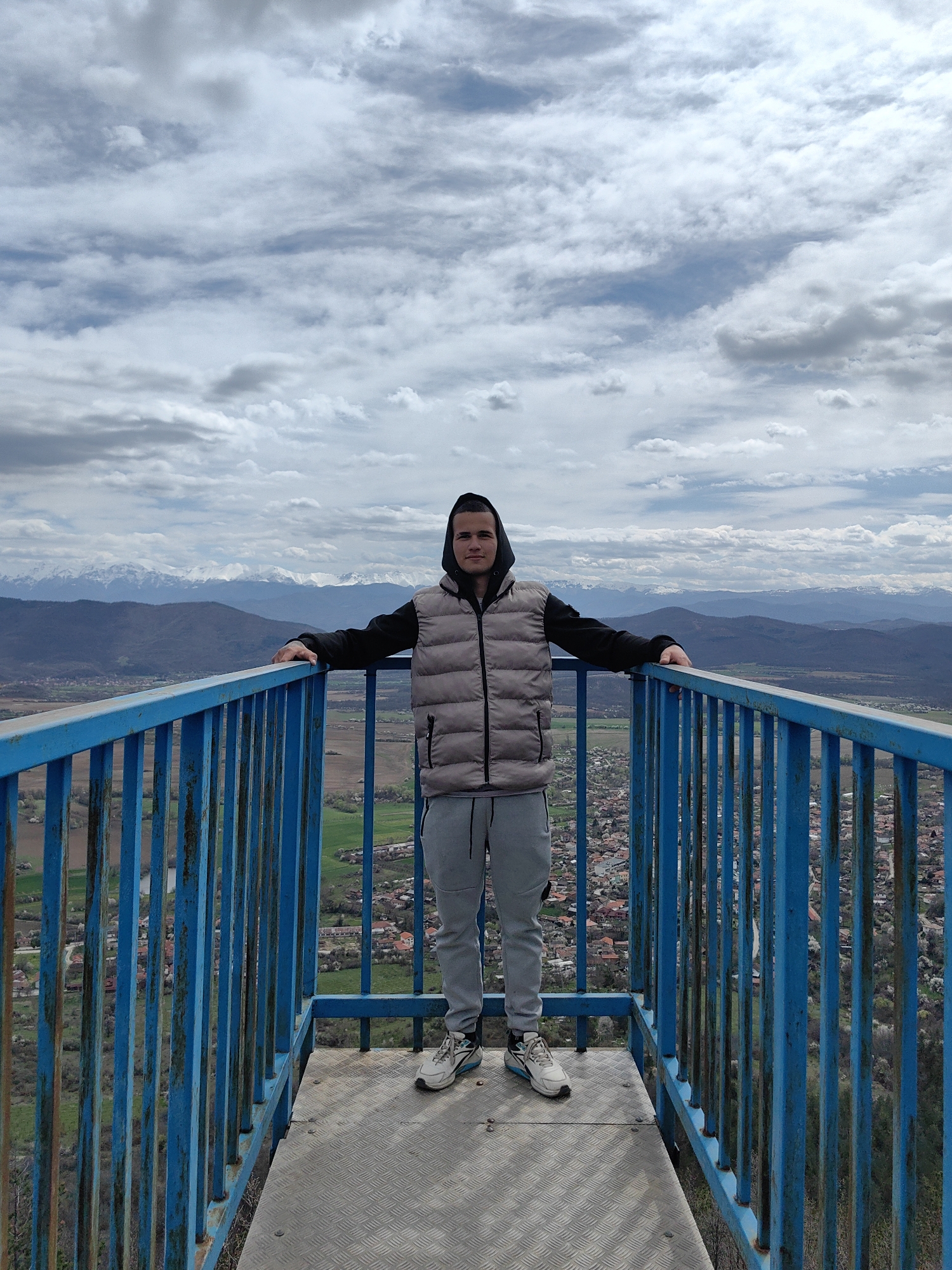 A full shot captures a young man standing on a blue metal observation deck, likely in the city of Vrabevo, Bulgaria. He faces the camera with his arms outstretched, resting his hands on the railing. He wears a black hooded sweatshirt, a gray quilted vest, gray sweatpants, and sneakers. The deck is made of a diamond-plated metal and surrounded by a bright blue metal railing.
The background reveals a wide, panoramic view. Below the deck, a town with red-roofed buildings is visible, interspersed with green fields and patches of trees. Further in the distance, rolling hills and mountains fade into the horizon. Snow-capped peaks are visible on the distant mountain range. The sky is filled with gray and white clouds, suggesting an overcast day.
