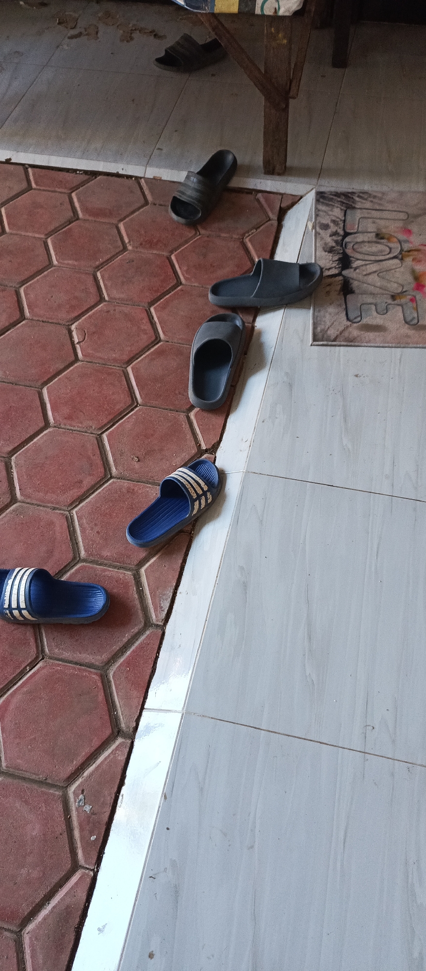 A collection of six pairs of flip-flops are scattered on a tiled floor. The floor is a mix of hexagonal red tiles and larger, rectangular light gray tiles. The flip-flops are of various colors and styles, including black, dark gray, and blue with white stripes. One pair of blue flip-flops with white stripes is particularly prominent in the foreground. The scene appears to be an interior space, possibly a home or a communal area, with a wooden table leg visible in the background. The lighting suggests it is daytime. There are no people or animals present in the image.