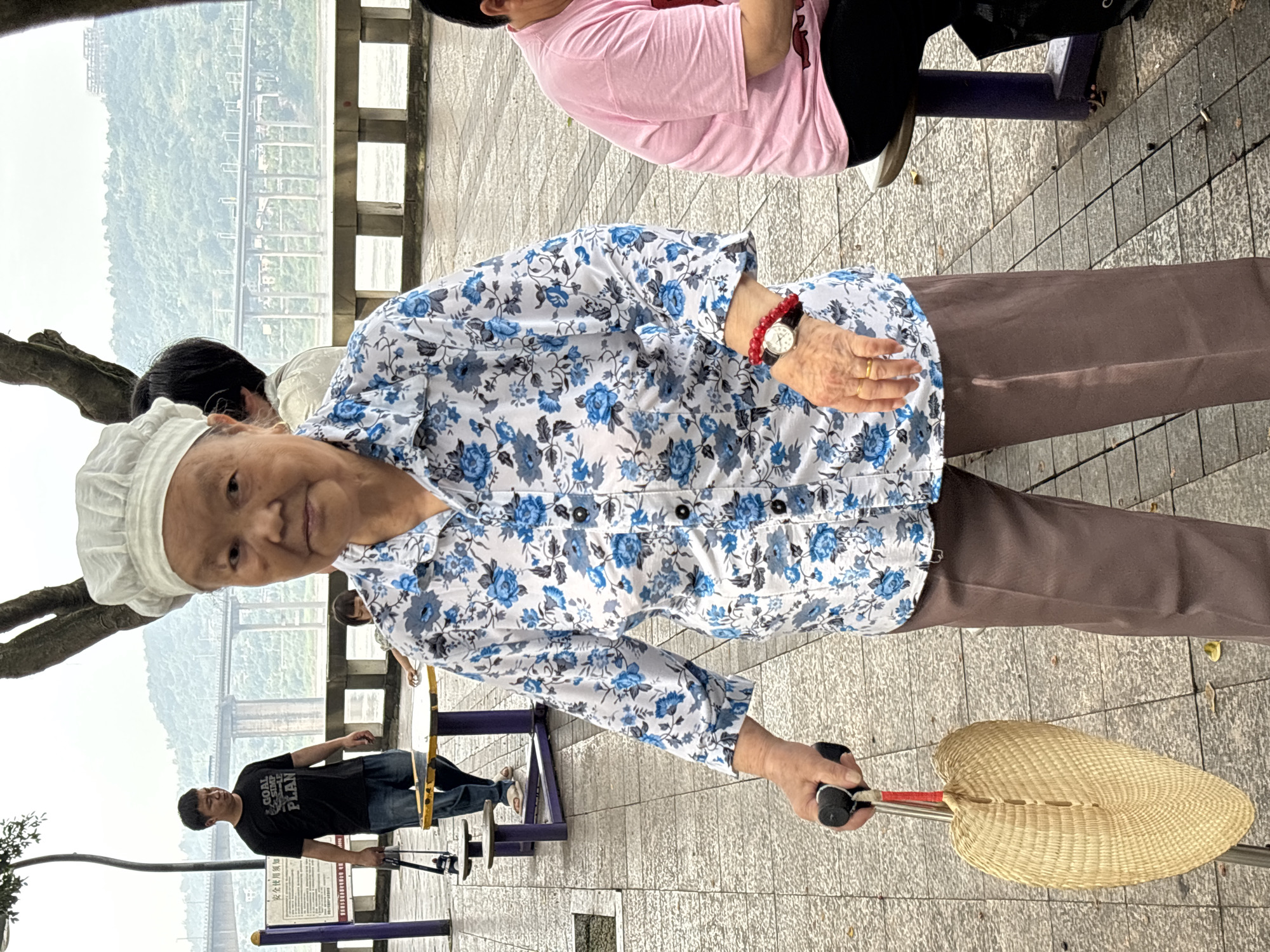 An elderly Asian woman wearing a white chef's hat and a blue floral print shirt stands on a paved walkway, holding a cane with a woven fan attached. She has a gold ring on her left ring finger and a red beaded bracelet on her left wrist with a watch. To her right, a person in a pink shirt sits on a bench. In the background, a man in a black t-shirt and jeans walks past a table and benches. Further in the background, a bridge spans a body of water, and a lush, green hillside rises beyond it. This scene is located in Jiangjin City, China, specifically along the Jialing River.