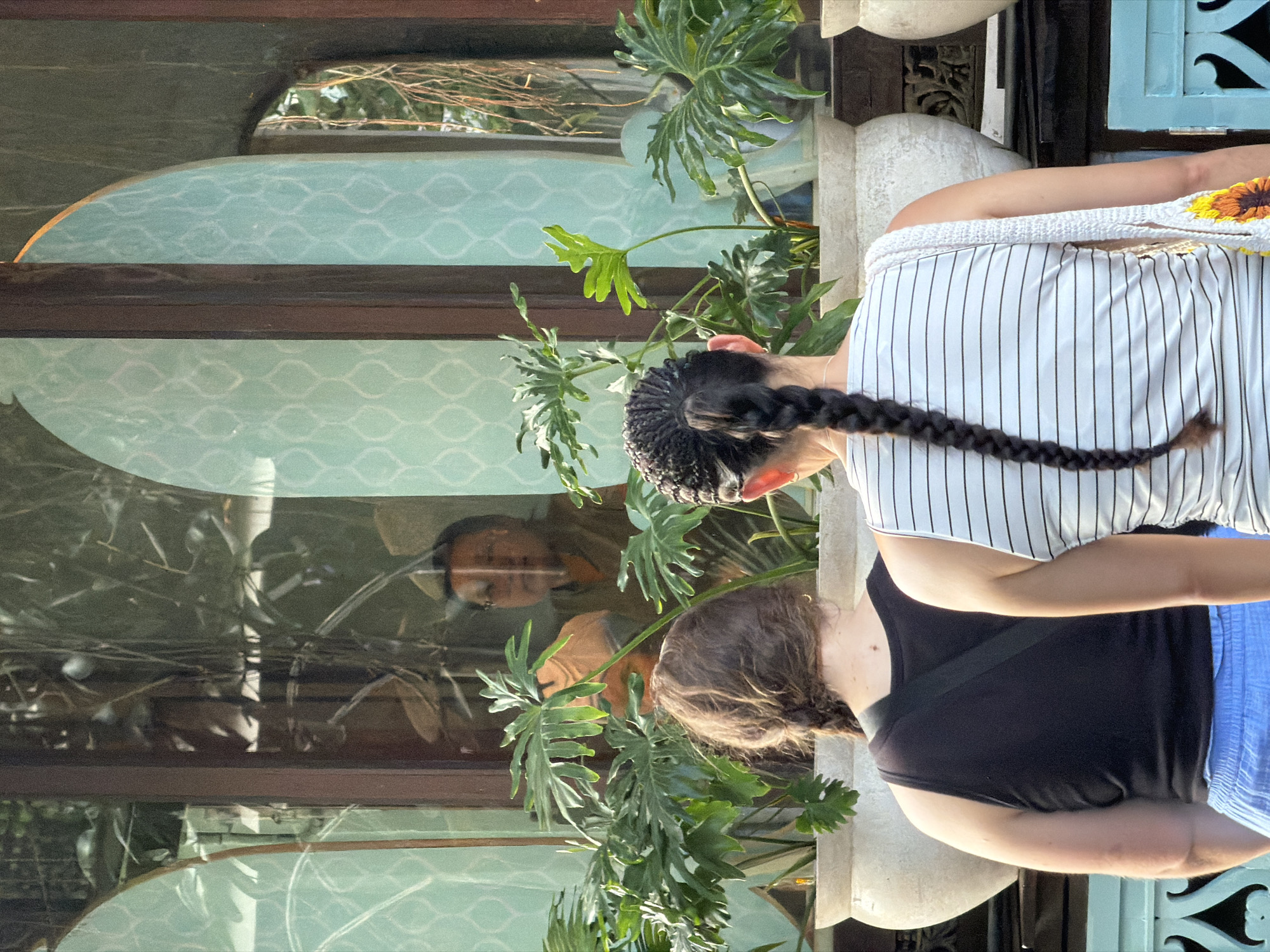 The image depicts two individuals standing with their backs to the camera, facing a large window. One person on the left has long, brown hair in a braid and is wearing a black tank top and blue shorts. The person on the right has black hair styled in a long braid and is wearing a white and black striped tank top and carrying a woven bag. Through the window, a reflection shows a person wearing glasses and an orange-brown garment, possibly a monk, seated indoors amidst plants. The setting appears to be a garden or outdoor seating area, given the presence of potted plants and natural light. The window itself features decorative patterns. While the exact location is not explicitly stated in the image, the prompt specifies "Ubud District, Indonesia."