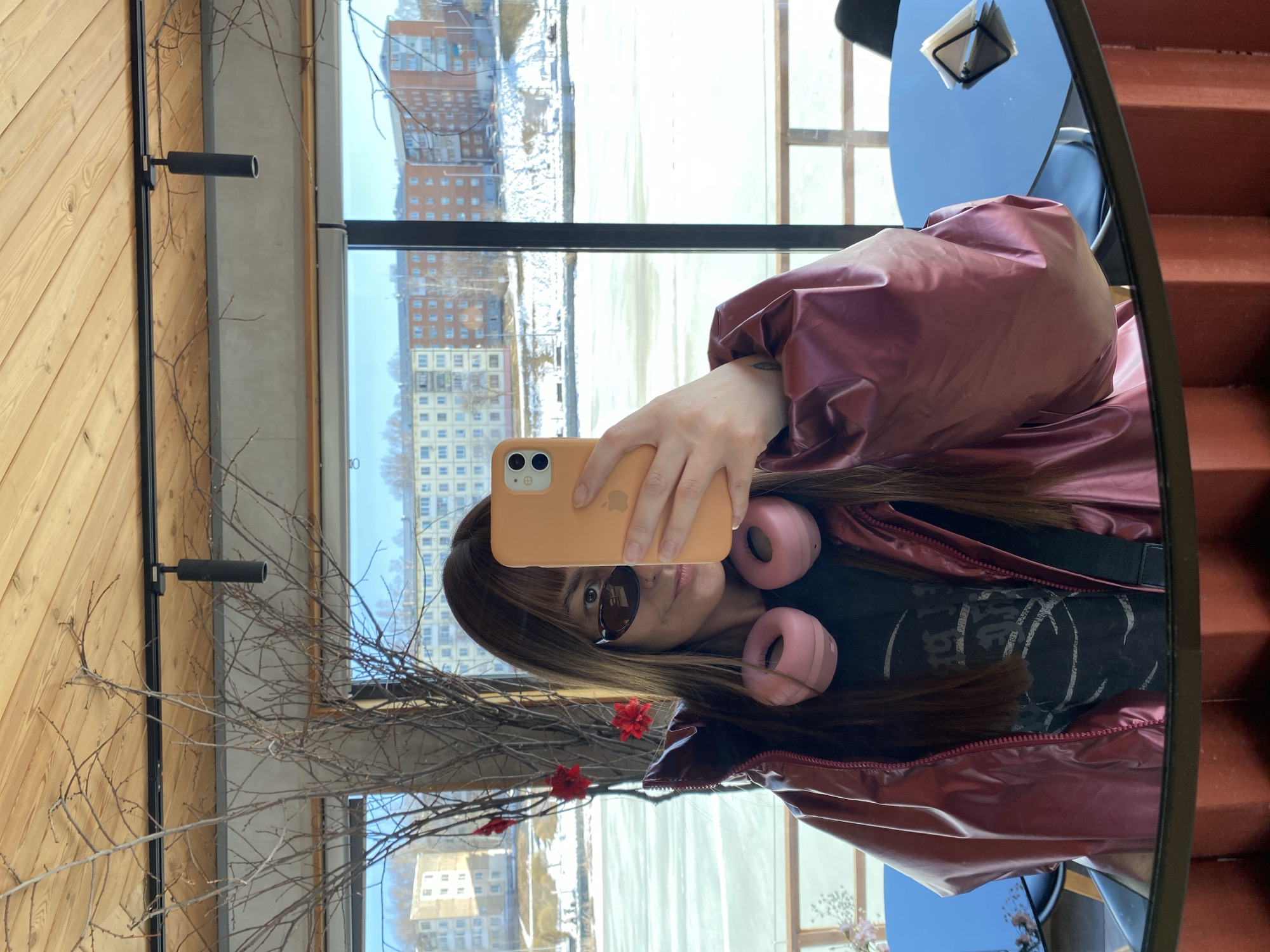 A woman is taking a selfie in a mirror, seemingly indoors. She is holding an orange phone, wearing sunglasses, and has pink headphones around her neck. She is wearing a shiny burgundy jacket. The reflection in the mirror shows a building exterior, possibly from a window, with a river or body of water visible. Other objects are visible such as the inside of a cafe/restaurant in which the woman sits. Based on the surrounding architectural elements and the details visible through the window, it's probable that the location is in Votkinsk, Russia.