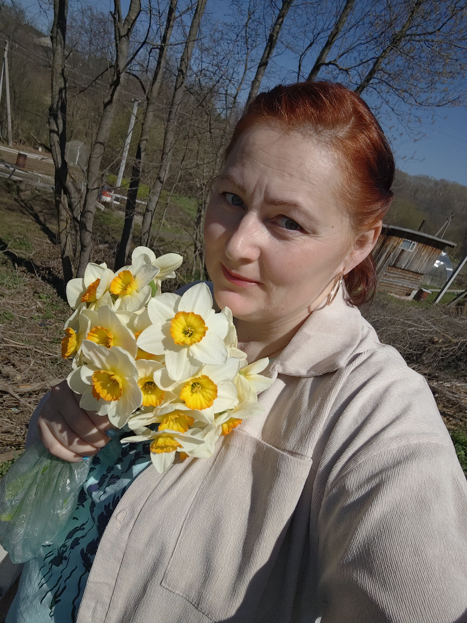 A red-haired woman holds a bouquet of white and yellow daffodils. The background consists of leafless trees and a partly visible wooden structure. The setting appears to be outdoors in daylight. The location is likely the city of Фоки, Russia, based on the provided context.