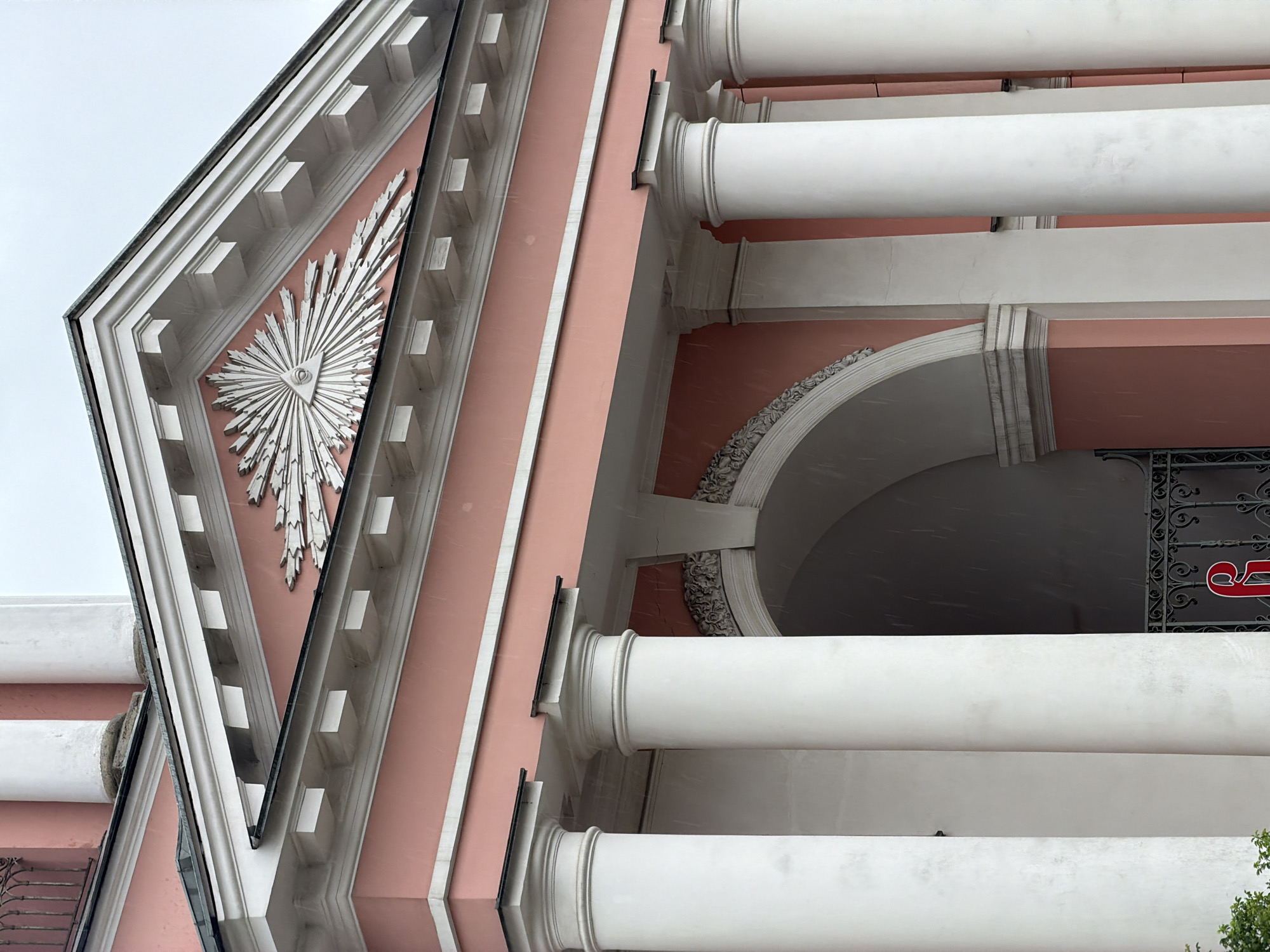 The image captures a portion of the facade of the Mikhailovsky Manege building in Saint Petersburg, Russia. The building's architectural details are prominently featured, including white columns, a pink exterior, and a triangular pediment with an ornate, sunburst-like emblem. The building's construction style includes classical elements like arches and decorative moldings. The sky is overcast. The image is taken from a low angle, looking up at the building.
