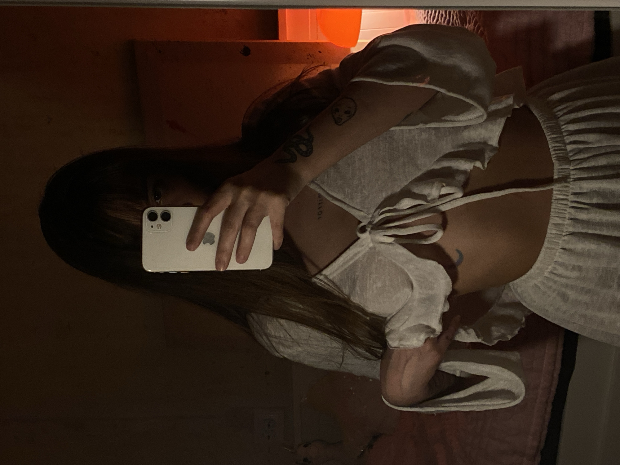 A person is taking a picture of themselves in a mirror. They are holding a white iPhone in their left hand, covering part of their face. The person has dark hair with bangs. They are wearing a white dress with a tie at the back. A skull and a snake tattoo are visible on the person's left arm. A moon tattoo is on the person's back. The location is unknown, and there is no information in the image to suggest that the photograph was taken in Votkinsk, Russia. The background is dark and blurry, with indistinct shapes of furniture and other objects.