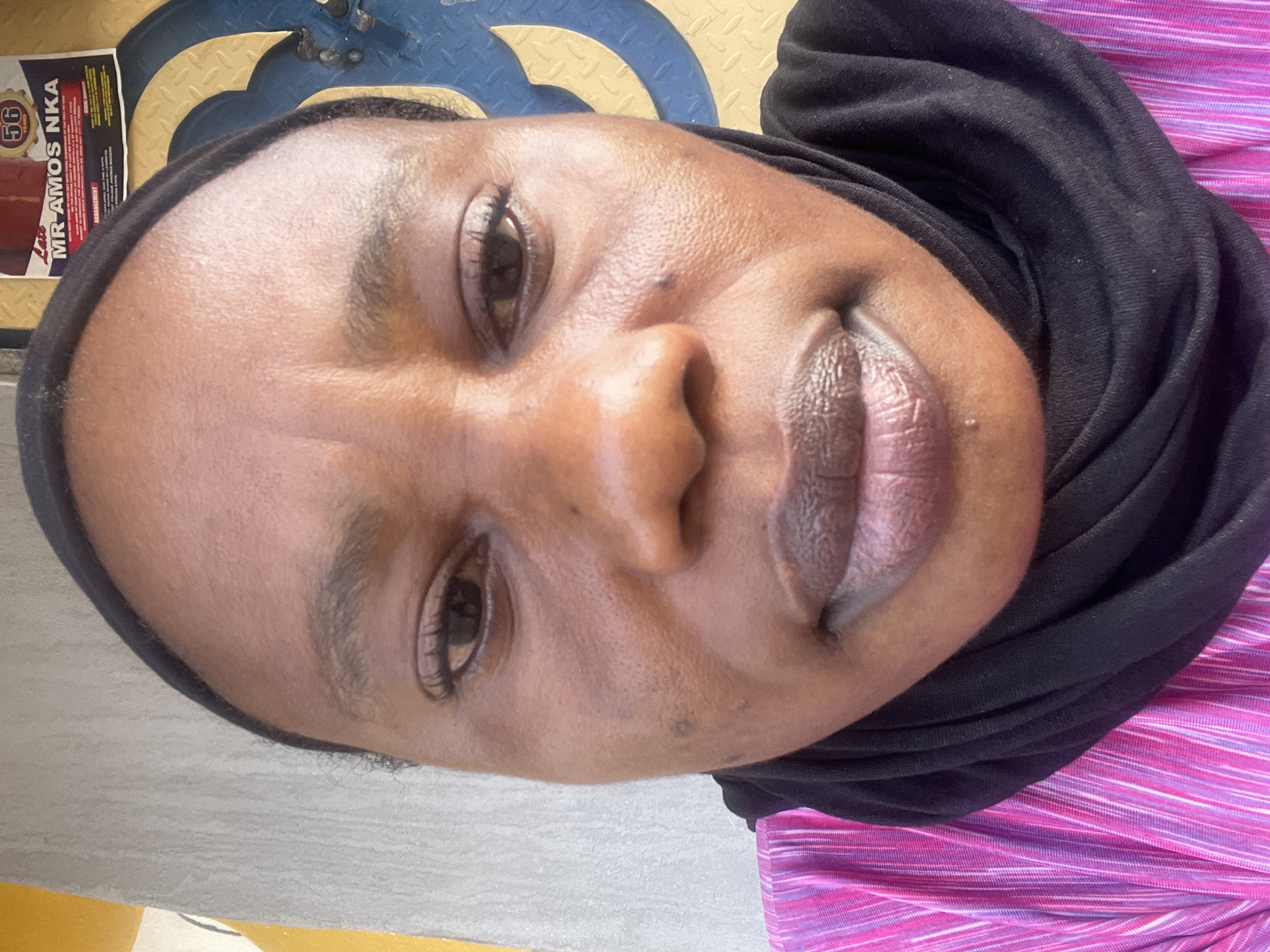 The image is a close-up selfie of a woman with brown skin, wearing a black hijab and a pink and white striped shirt. Her face is the main focus, with details such as her eyes, nose, and lips clearly visible. A sticker with "MR AMOS NKA" is on the top left corner of the image. The background is slightly blurred, revealing a blue and yellow patterned surface. There is no indication of a specific location within Rido, Nigeria.