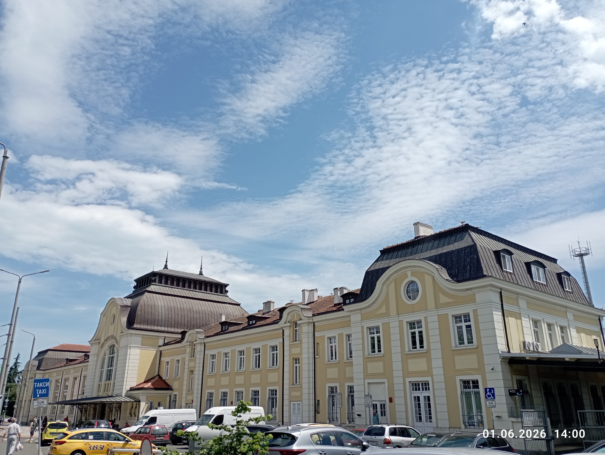 The image shows a section of the Plovdiv Railway Station, in Plovdiv, Bulgaria. The building is a long, low structure of two or three stories, painted yellow and white. It is topped by a dark roof with rounded dormers on the right. There are several cars parked in front of the building, and a white van. On the left, there is a yellow taxi cab with "TAXI" written on it. In the background, there are more trees and buildings, as well as a tall antenna. The sky is blue with scattered white clouds. In the lower right corner, there is the date "01.06.2026 14:00". There are also a few people on the left, walking in front of the building.
