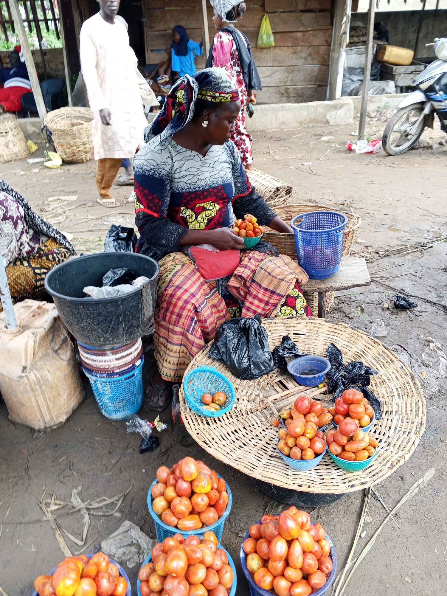 This image depicts a busy market scene, likely in Lokoja, Nigeria, characterized by a vibrant display of produce and human activity. In the foreground, a woman wearing a colorful headwrap and patterned dress is seated, attending to a selection of tomatoes arranged in woven baskets and blue plastic containers. Her hands are visible, holding a small green bowl filled with smaller fruits, possibly plum tomatoes or similar produce. Adjacent to her are various baskets, some empty and others containing more tomatoes and what appear to be some onions. A large black plastic bucket, filled with what looks like trash bags, is stacked on top of several colorful plastic baskets. To the left, a large woven basket sits on the ground, and a pale beige sack leans against a metal pole.
In the middle ground, a man dressed in a long white garment and brown trousers stands, facing away from the camera, his attention seemingly directed towards something beyond the frame. Further back, a young girl in a bright blue headscarf and dress is visible, standing near a wooden structure. Other market stalls and a motorcycle are also present in the background, suggesting a bustling commercial environment. The ground is a mixture of dirt and debris, indicating an outdoor, possibly informal, marketplace. The overall impression is one of everyday commerce in a Nigerian city, with the focus on fresh produce and the people who sell and buy it.