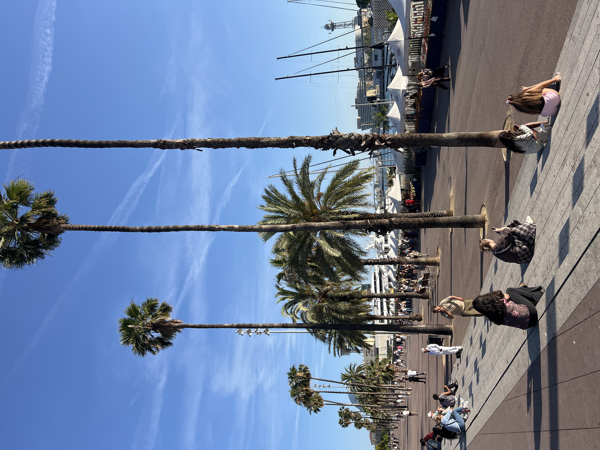 This image depicts a wide, sunny promenade lined with numerous tall palm trees, located in the Port Vell area of Barcelona, Spain, specifically along the Moll de la Fusta or an adjacent waterfront section. The scene is illuminated by bright sunlight under a clear blue sky with several faint white contrails.
In the foreground and midground, a diverse group of people is present. Several individuals are sitting on a raised, grey-tiled platform that borders the promenade. On the left, a person in a light blue top and jeans is seated. In the center, a woman with long dark hair, wearing a floral top and dark pants, sits facing away from the camera. To her right, another woman in a plaid skirt and dark top is seated, engrossed in a mobile phone. Further right, two more individuals are seated: one in a striped top and dark shorts, and another in pink shorts and a dark top, also seemingly engaged with phones. A man in a light tan shirt and dark pants stands to the left of these seated individuals, appearing to hold something small to his mouth. Another man in a white suit stands further back, facing away.
The wide promenade is paved with distinct sections of reddish-brown and grey tiles. Many people are seen walking in various directions along the promenade in the midground and background, populating the area.
On the right side of the image, beyond the palm trees, a body of water is visible, with numerous sailboats and yachts docked. Their tall masts rise against the skyline. Along the waterfront, a series of identical white market or event tents are set up in a row. In the distant background, several urban buildings are visible, including a prominent structure with a pointed top, characteristic of Barcelona's port architecture (e.g., near the World Trade Center or a cable car tower). Tall, slender streetlights are interspersed among the palm trees. Strong shadows are cast across the promenade from the trees and people, indicating the time of day is either morning or late afternoon.