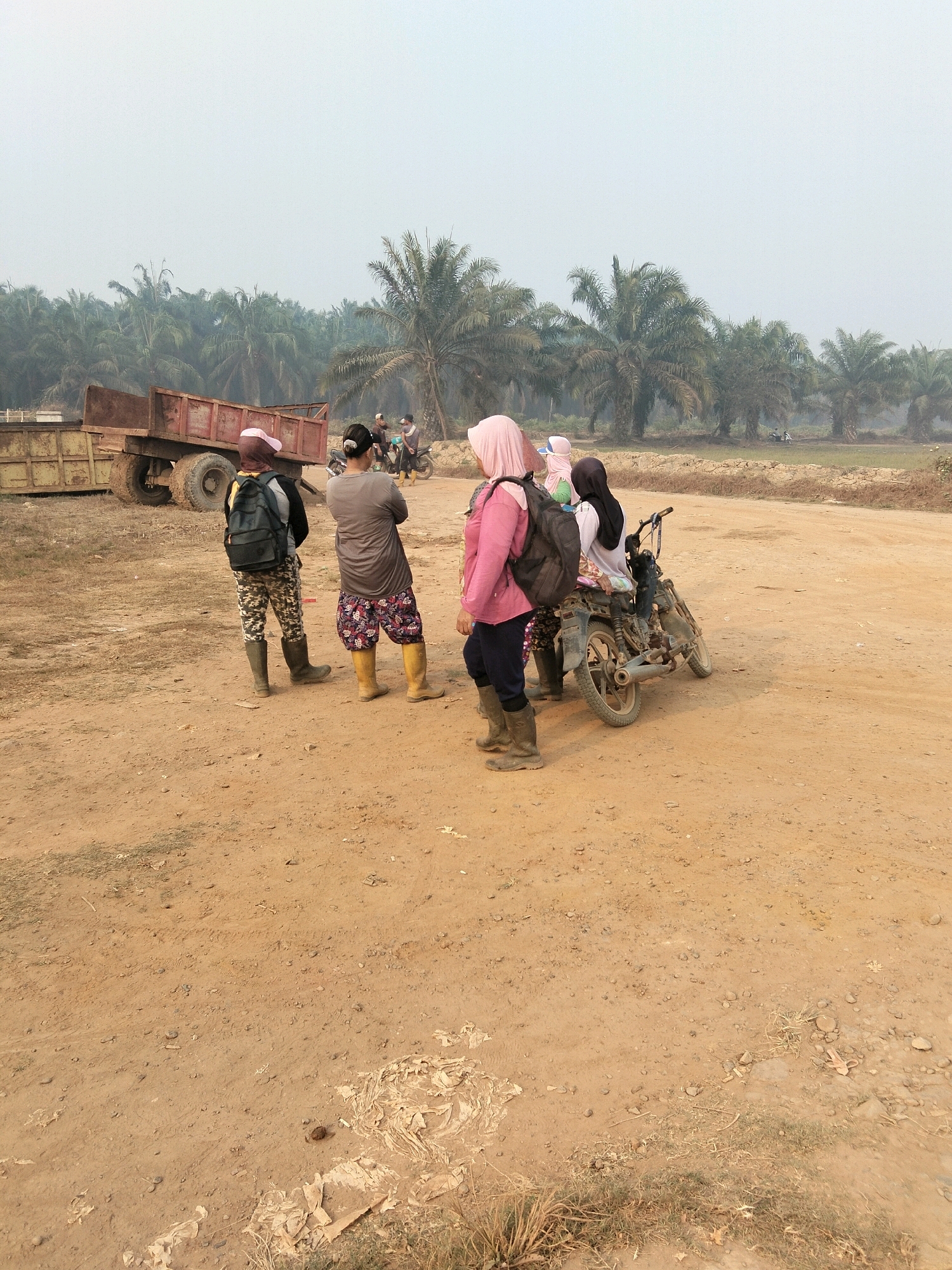 In Teluk Tenggirik, Indonesia, a group of five individuals are gathered on a dusty, unpaved road, with a plantation of palm trees in the background. Two individuals, a man and a woman, are standing near a rusty, old truck. Another woman, wearing a pink headscarf and a backpack, is standing by a motorcycle. Two more people are on the motorcycle, one of them wearing a pink headscarf. The individuals appear to be in a rural setting, possibly working in the plantation or taking a break. The atmosphere is hazy, suggesting a dry or polluted environment.