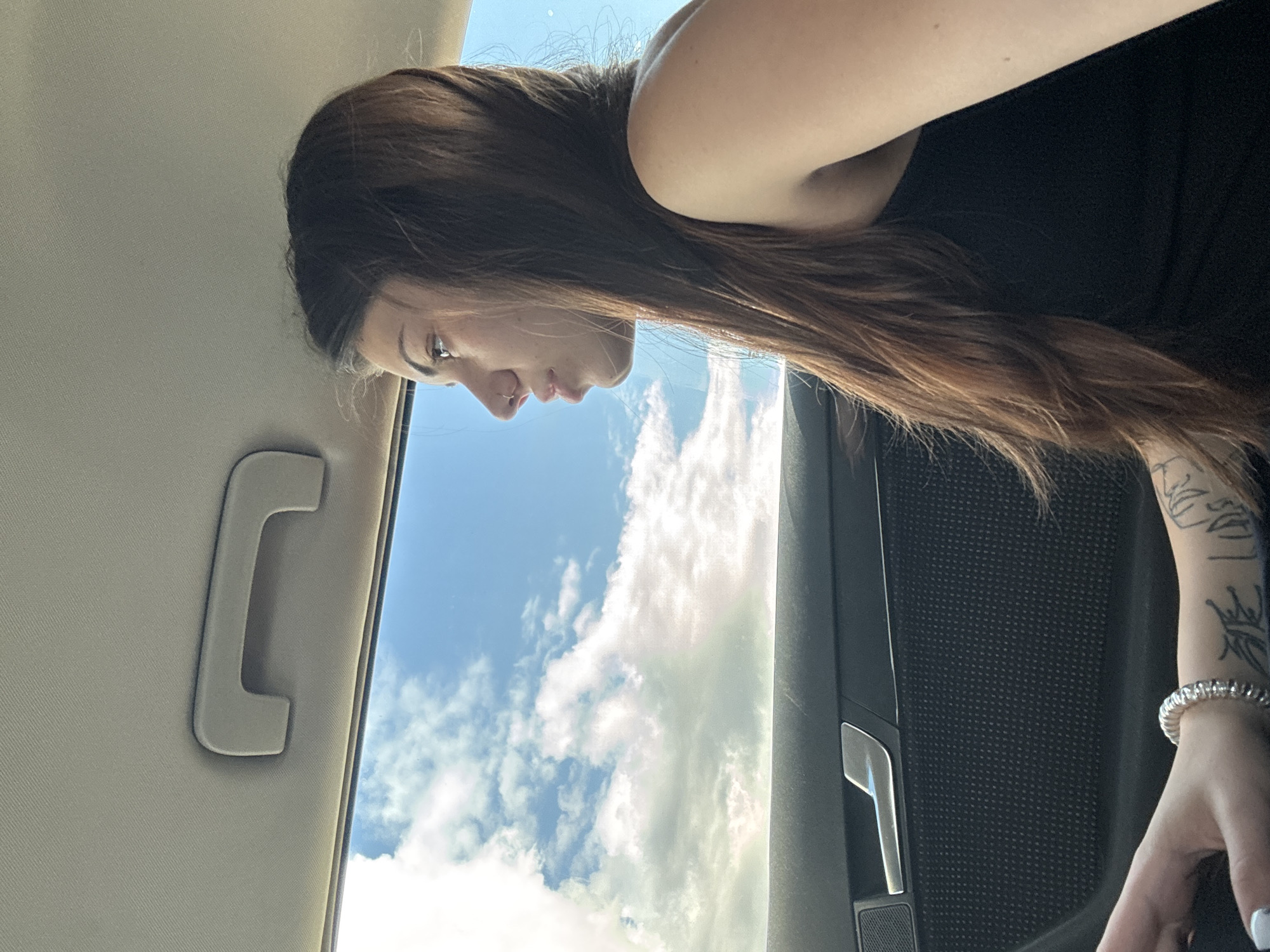 A close-up, rotated shot captures a person with long, dark brown hair from the shoulders up, positioned within a vehicle's interior. The person is seen in profile, looking upwards and slightly to the right, with their face illuminated by natural light. Their left arm is visible, adorned with a black tattoo featuring crown-like designs and a silver beaded bracelet. They wear a black top.
The setting appears to be the inside of a car, with a light-colored headliner and a gray grab handle visible on the left. A dark door panel with a textured surface and a metallic door handle is seen on the right. Through what seems to be a sunroof or a large car window, a bright blue sky filled with white, fluffy clouds is prominently featured, suggesting a clear and sunny day. The lighting indicates it is daytime. The overall impression is a moment of observation or contemplation while traveling in Mramor, Bulgaria. No visible text is present in the image.