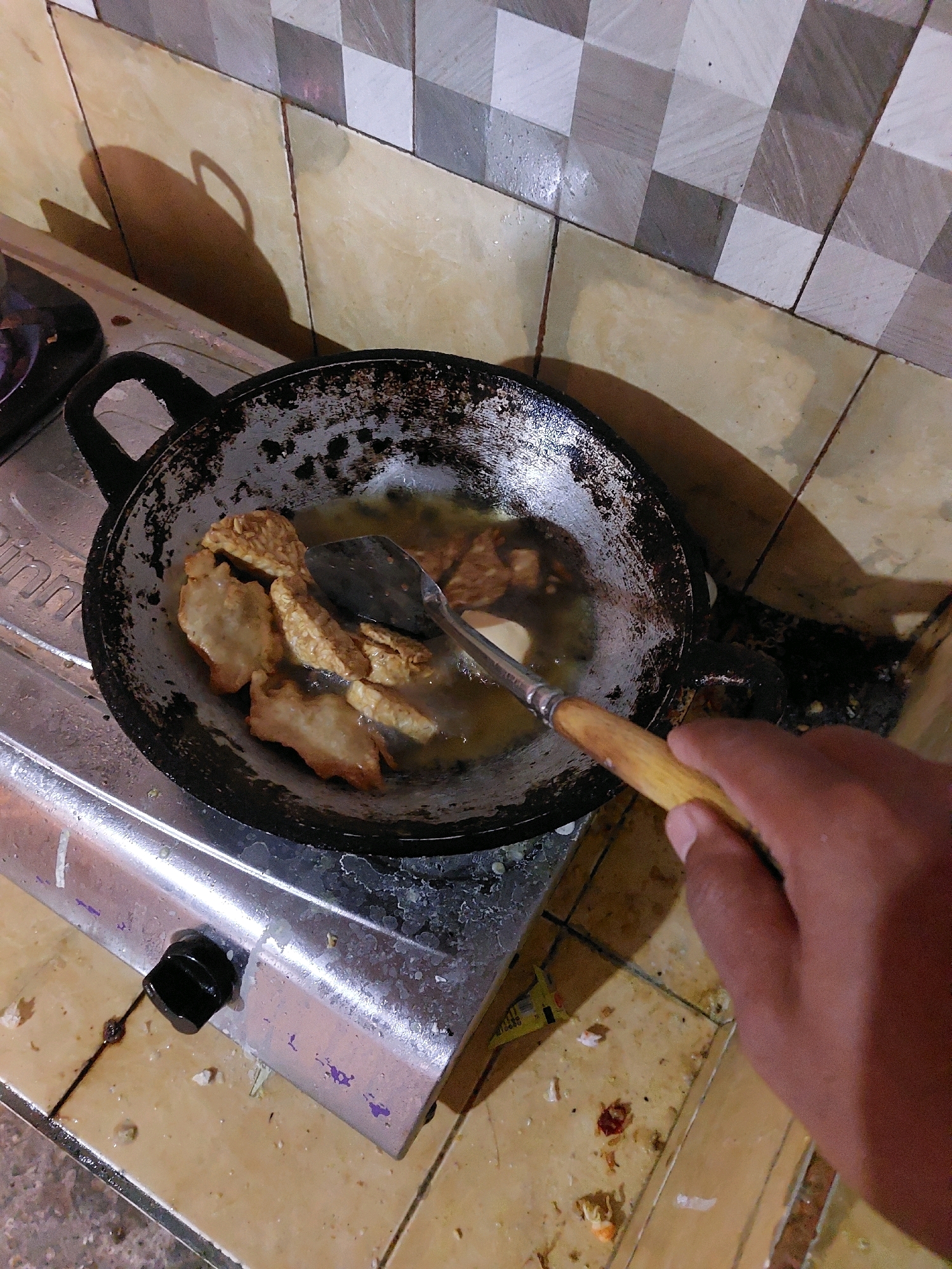 In the city of SARANGAN, Indonesia, a close-up, overhead shot captures a cooking scene on a stovetop. The focus is on a well-used black frying pan filled with hot oil, in which several pieces of what appear to be fried tempeh are cooking. A metal spatula with a wooden handle is submerged in the oil, being used to turn the food. A person's hand, with brown skin tone, is visible on the right side of the frame, gripping the spatula handle. The frying pan rests on a silver gas stove, with one black knob visible on the front. The background features a wall tiled with square patterns of light yellow and grey, with a patterned backsplash above. The overall environment appears to be a domestic kitchen, with some evidence of splatters and cooking residue on the stovetop and surrounding tiles.
