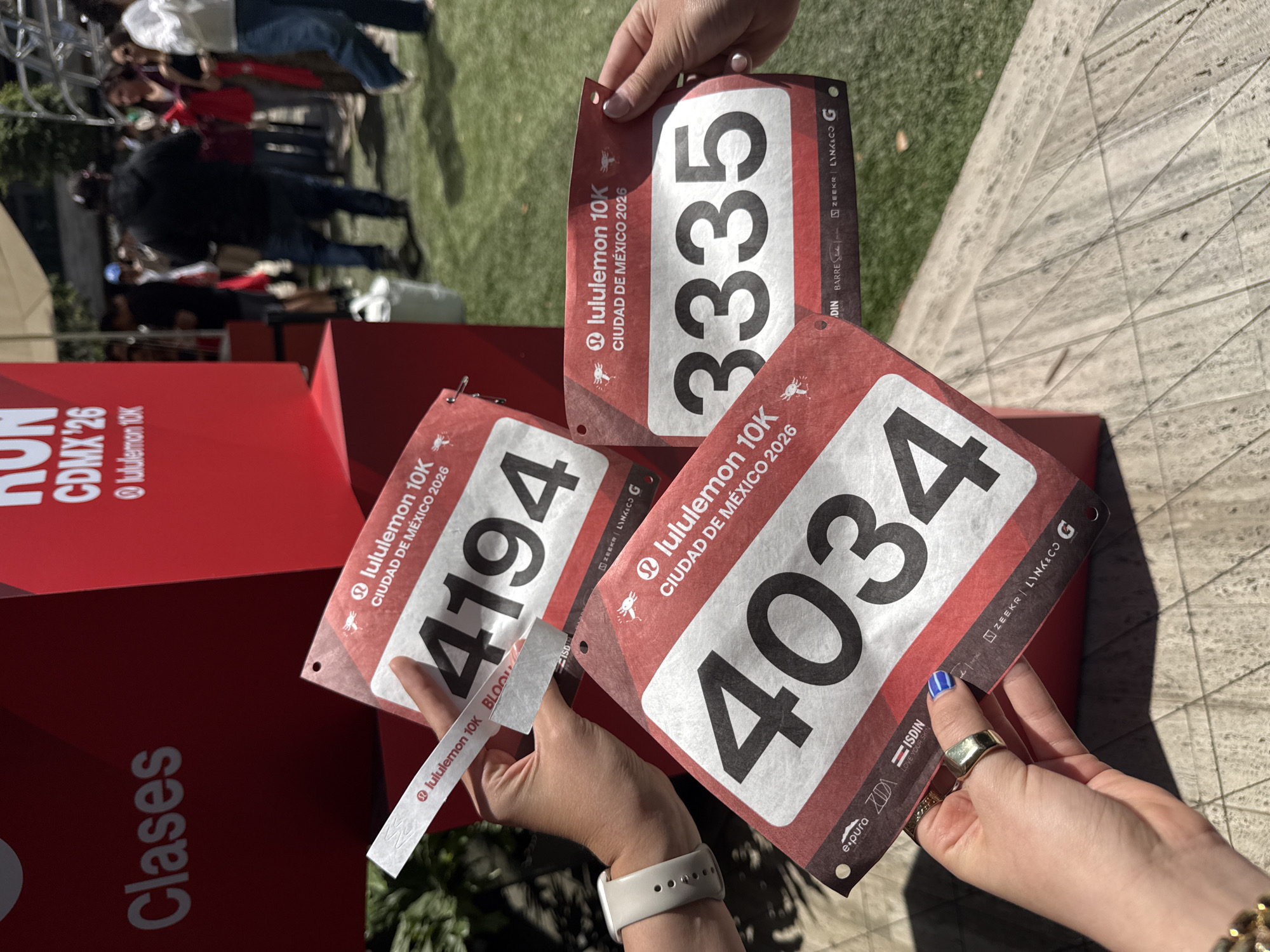 Here is a description of the image:
The image shows three race bibs being held up, likely for a "lululemon 10K" event in Mexico City in 2026. The bib numbers are 4194, 3335, and 4034. The bibs feature the lululemon logo and mention sponsors such as ZEEKR, LINK&CO, ISDIN, and e.pura. A wristband that says lululemon 10k is being held with the bib that says 4194. In the background is a red display that has the word "Clases" on it. People can be seen milling about in the background, indicating the event is taking place outdoors.