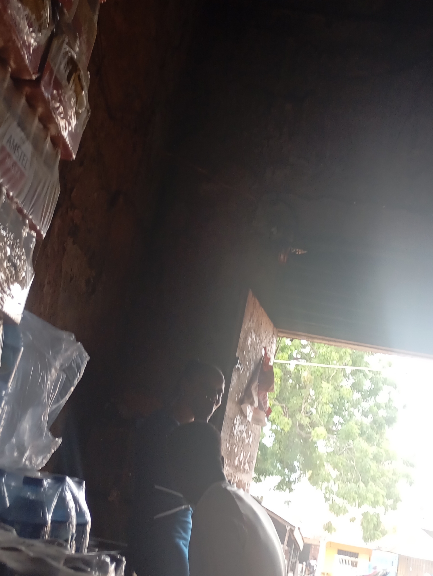 The image depicts an interior scene of what appears to be a small shop or storage area, possibly in a developing urban environment given the rustic construction visible. In the foreground, stacked packages, some wrapped in plastic, are partially visible. The packaging on the left side of the image contains bottles, some of which appear to be water bottles based on the clear plastic and labels. One package on the far left displays the word "AMSTEL".
The middle ground features two individuals. One, partially obscured by shadow, is wearing a dark shirt and appears to be interacting with the other person. The second individual, also in shadow, is facing away from the camera and is more clearly defined. They are wearing a dark garment and blue pants. A large, light-colored, curved object, possibly a bag or a piece of equipment, is positioned between the two individuals, obscuring some of the lower half of their bodies.
The background reveals an open doorway or window leading to an outdoor area. Bright daylight illuminates a lush green tree and some buildings in the distance. The interior walls are rough, made of what looks like unplastered brick or mud, suggesting a basic construction. The overall lighting is dim inside, with a strong contrast to the bright exterior. The image quality is low, with significant noise and blur, making precise identification of details challenging.