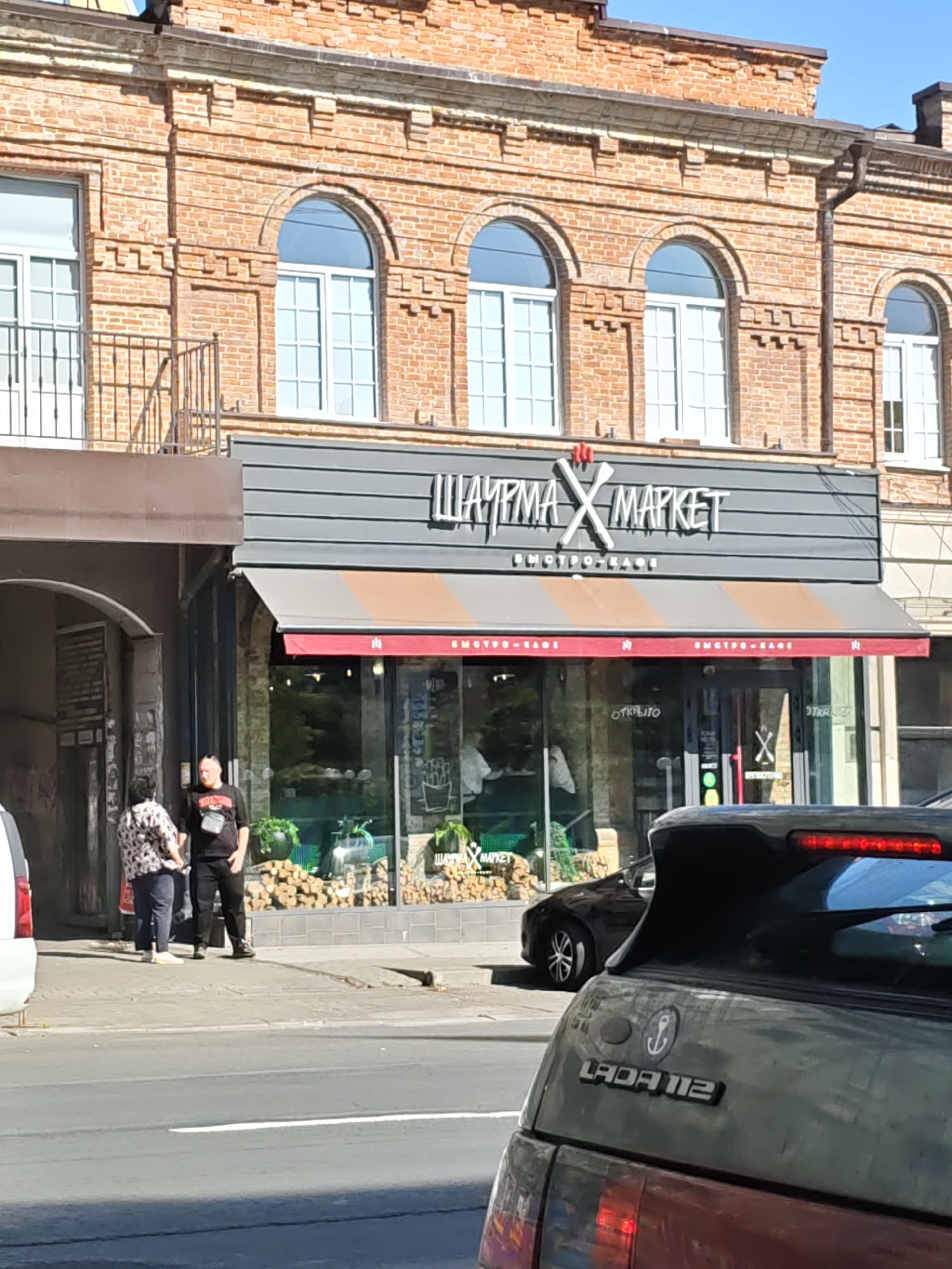 The image shows a street view in Rostov-on-Don, Russia, featuring the facade of a brick building housing a food establishment called "ШАУРМА Х МАРКЕТ" (SHAURMA X MARKET). The building has arched windows on the upper floor and a storefront with a striped awning on the lower level. Two individuals are standing on the sidewalk in front of an archway to the left of the market. One person, appearing to be male, is wearing a black t-shirt and black pants, with a crossbody bag. The other person, appearing to be female, is wearing a patterned shirt and blue pants. A dark-colored car is parked in front of the market's storefront. In the foreground, the rear of a dark-colored Lada 112 car is visible, with its spoiler illuminated by brake lights. The scene is illuminated by daylight, suggesting it was taken during the day.