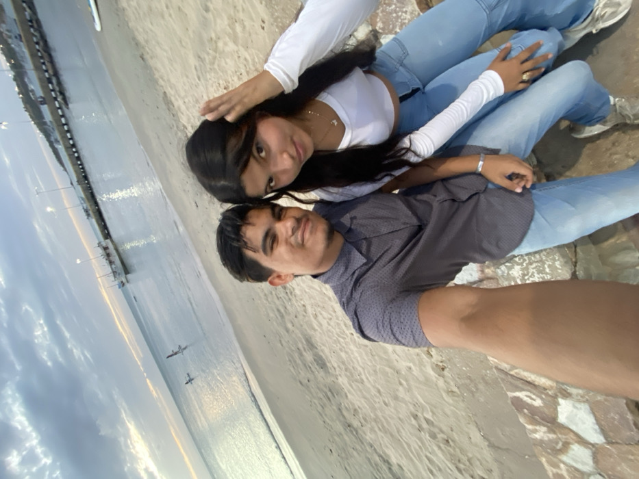 A young couple poses for a selfie on a beach. The woman, with long dark hair, wears a white long-sleeved crop top and light jeans. Her hand rests on her head. The man, wearing a dark patterned shirt and light jeans, smiles at the camera. The ocean and a pier stretch into the background under a cloudy sky. The pair sits on a rocky area near the sand.