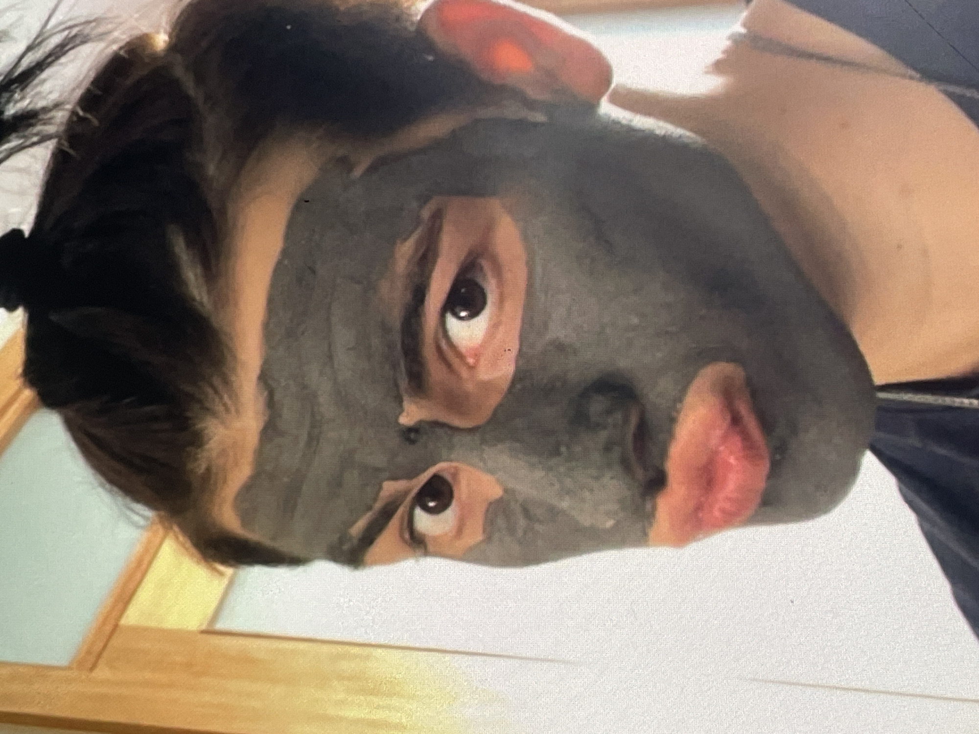 The image is a close-up, portrait-style photograph of a person wearing a dark grey face mask that covers their entire face except for their eyes and lips. The person has dark hair pulled up in a bun on top of their head. Their lips are painted a bright pink or coral color and are slightly parted, with the upper lip appearing more prominent. One eye is clearly visible, looking directly at the camera, and the other is partially obscured by the mask. The person's skin tone is fair. The background consists of a blurred interior setting, with a wooden door frame and a light-colored wall visible to the left. The lighting is bright, causing a slight glow on the person's ear. There is no indication of the city of Harmanli, Bulgaria, in the image.