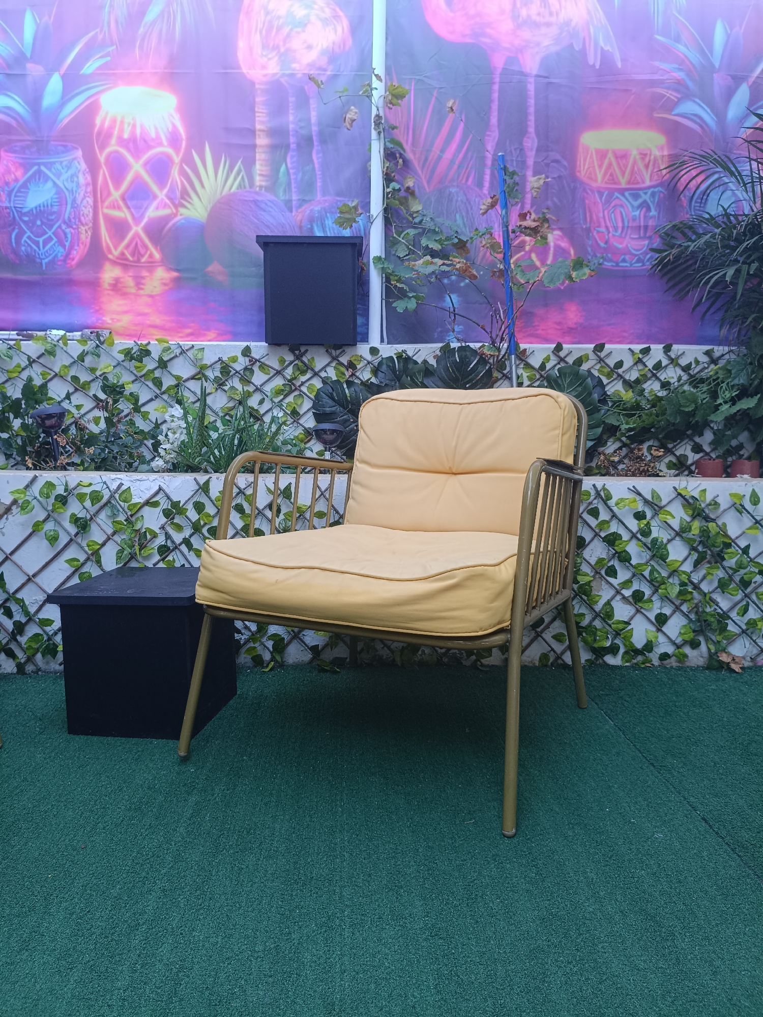 In the city of Rafelbunyol, Spain, an outdoor seating area is depicted. The scene features a yellow cushioned chair with a gold-colored metal frame positioned on a green carpeted surface. To the left of the chair is a black rectangular box. Behind the chair, a white lattice fence is visible, adorned with green vines. Beyond the fence, a vibrant backdrop is displayed, featuring stylized illustrations of pineapples and flamingos in neon colors. The backdrop is illuminated, creating a colorful and somewhat surreal atmosphere. The lighting suggests it is either evening or an indoor space with simulated outdoor lighting. There are no people present in the image.