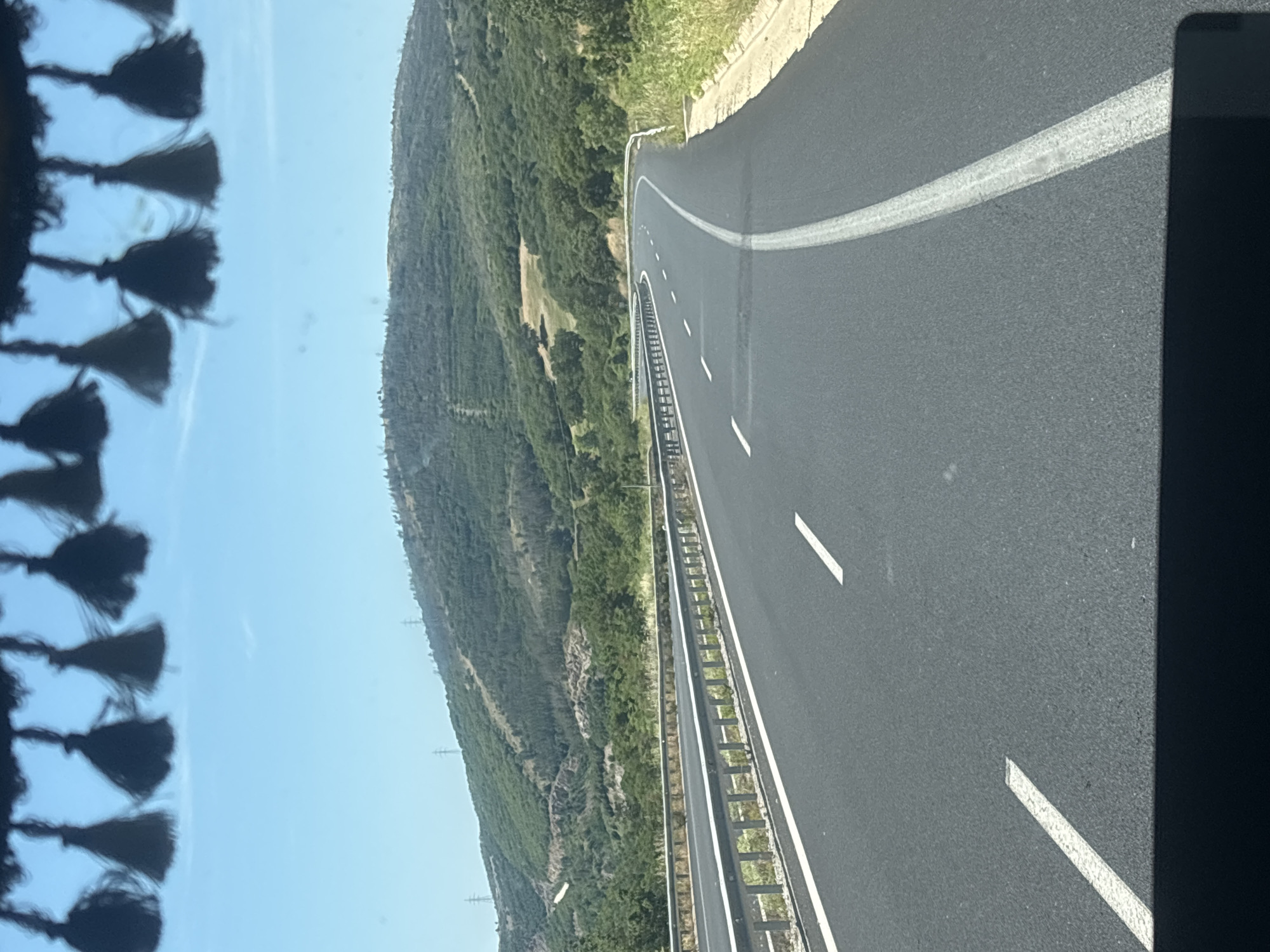The image depicts a view from inside a vehicle looking out at a highway winding through a hilly, vegetated landscape. The road is asphalt with white lane markings. A metal guardrail runs along the left side of the road. In the background, a green, tree-covered hill rises, with power lines visible on its slopes. The sky is clear and blue. The foreground shows the interior of the vehicle, with decorative tassels hanging from the top edge of the windshield and a dark dashboard at the very bottom. The scene suggests a sunny day in a rural or semi-rural area. The location is specified as the city of Avra, Greece.