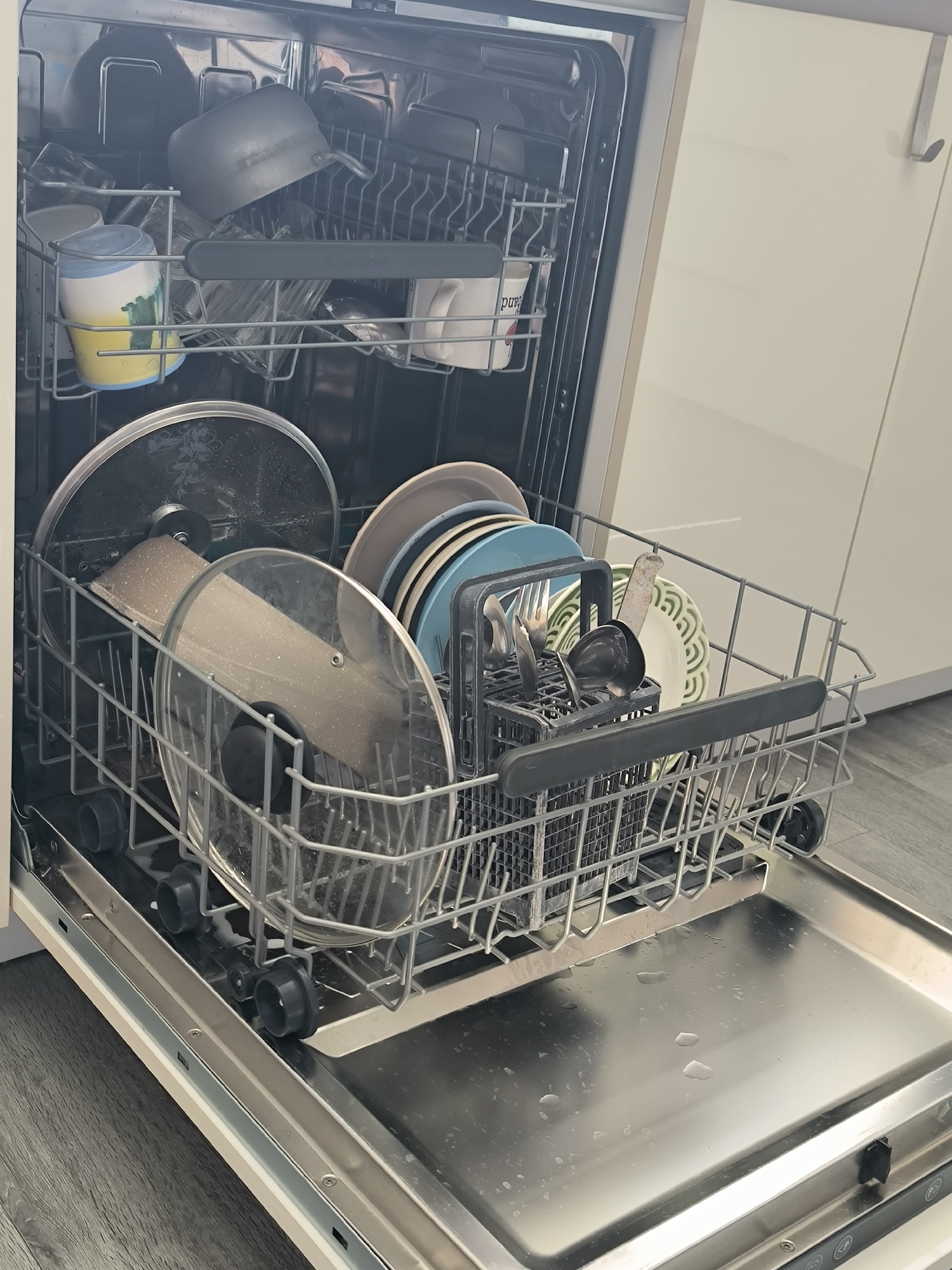 The image shows the interior of a dishwasher in an open state, located in the city of Oktyabrsky, Russia. The dishwasher is filled with various dishes, including pots, pans, lids, plates, and cutlery. The upper rack holds several items, including what appears to be a pot or saucepan, and some glasses or cups. The lower rack is more densely packed with dishes. Visible items include two large pot lids, a stack of plates of different sizes and colors, and a cutlery basket containing forks and spoons. The dishwasher is positioned in a kitchen setting, with the exterior of a white cabinet visible to the right of the appliance. The floor beneath the dishwasher is a grey wood-grain laminate. The lighting suggests an indoor environment, likely during the daytime.