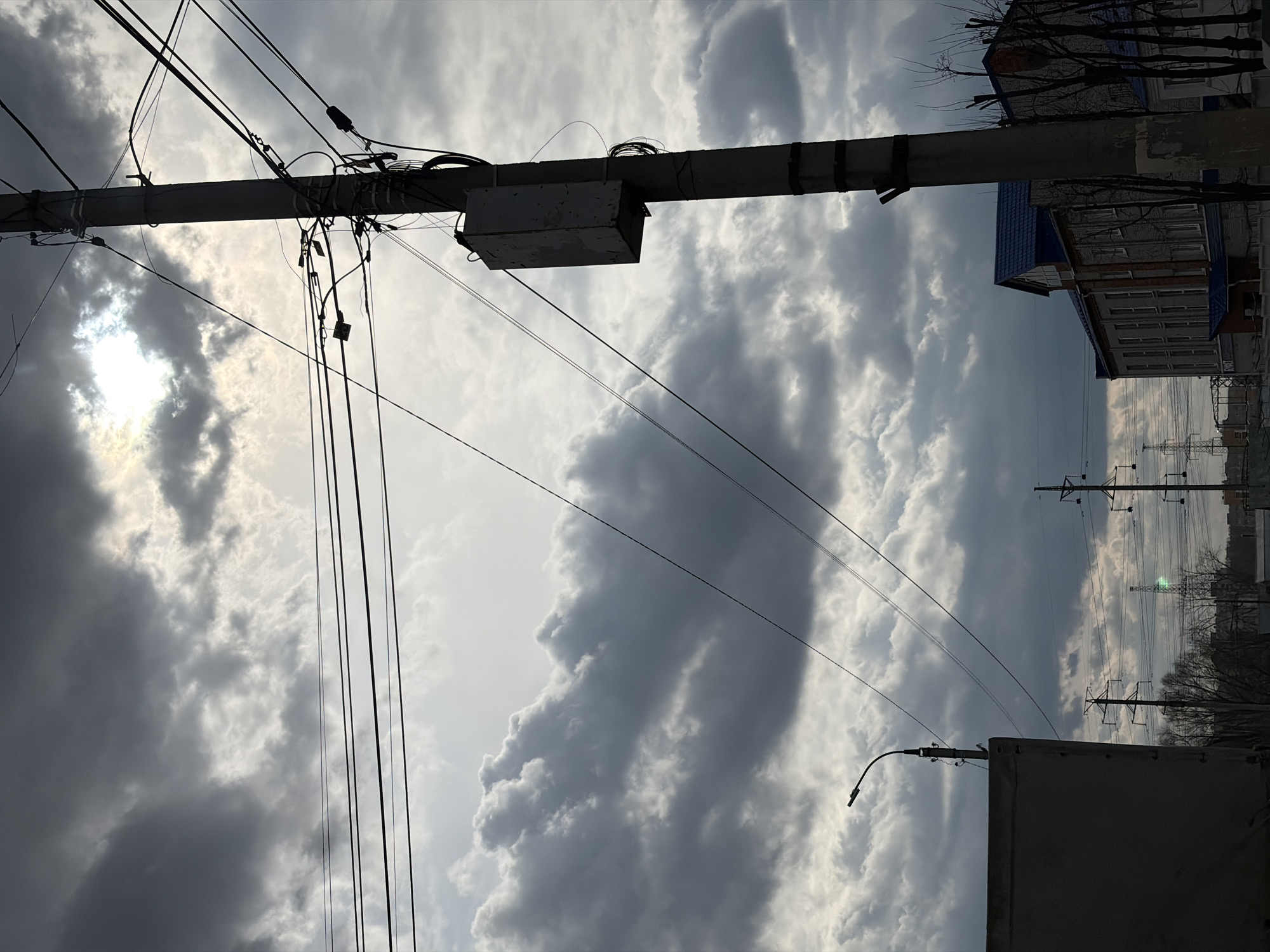 An outdoor, low-angle photograph captures a dense network of utility wires and cables against a cloudy sky in Chaikovsky, Russia. The primary subject is a dark, silhouetted utility pole running horizontally across the upper middle of the frame, laden with numerous black wires, insulators, and a rectangular utility box. The sun, partially obscured by bright clouds, creates a strong halo effect and backlights the scene from the upper left, casting the foreground elements into deep shadow.
The sky is a prominent feature, dominated by a mix of bright white and dark grey cumulus clouds, suggesting dynamic weather conditions. In the mid-ground and background, more power lines extend horizontally across the frame. To the right, part of a building with a blue roof and multiple windows is visible, along with the bare branches of trees, indicating a season like winter or early spring. Further in the distance, additional utility poles or power line towers are silhouetted against the lighter parts of the sky, with a faint greenish glint in one area that could be a lens flare or distant light.
No human activity is depicted, and the scene appears static. The time of day is likely daytime, possibly late morning or afternoon, given the bright sun. There is no visible text in the image.