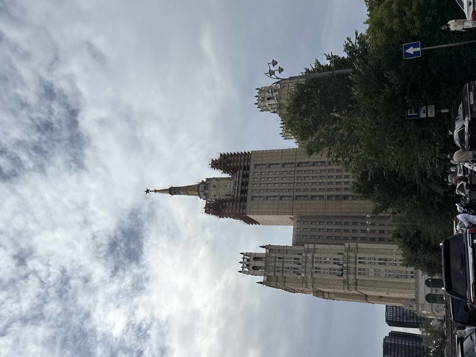 A low-angle, full shot captures the imposing Stalinist skyscraper, the Ministry of Foreign Affairs of Russia in Moscow, under a cloudy sky. The ornate, pale stone building dominates the frame, its elaborate architecture featuring multiple tiers, decorative spires, and a prominent red star atop its central spire. Scaffolding is visible on the upper sections of the central tower, suggesting ongoing construction or renovation. Flanking the central tower are two smaller, equally ornate towers, one to the left and one to the right. Lush green trees obscure the lower portion of the building on the right, partially obscuring a street lamp with multiple globes. In the foreground, several cars, including a black SUV, are parked, with a few pedestrians visible. Two dark, modern high-rise buildings are visible in the far left background, contrasting with the older architecture of the skyscraper. Two blue street signs are present: one indicating parking with times, and another with a white arrow pointing upwards. The sky is filled with dense, layered clouds, creating a somewhat overcast and dramatic atmosphere.