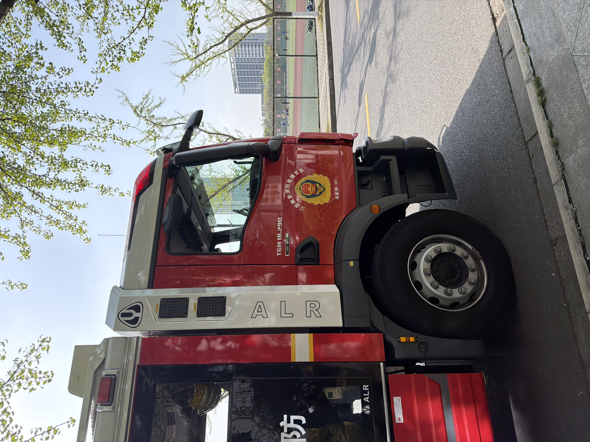 The image shows a red and white fire truck parked on a paved road, near a sidewalk. The side of the truck displays the letters "ALR" and a Chinese emblem with text indicating "Jiangsu Province Fire and Rescue Team" and "Baixia Lake-01", placing it in the Jiangning District of Nanjing, Jiangsu province, China. In the background, a sports field with a running track and a green fence is visible, with a tall building in the distance. A tree with green leaves is to the left of the fire truck. The overall scene suggests a sunny day.