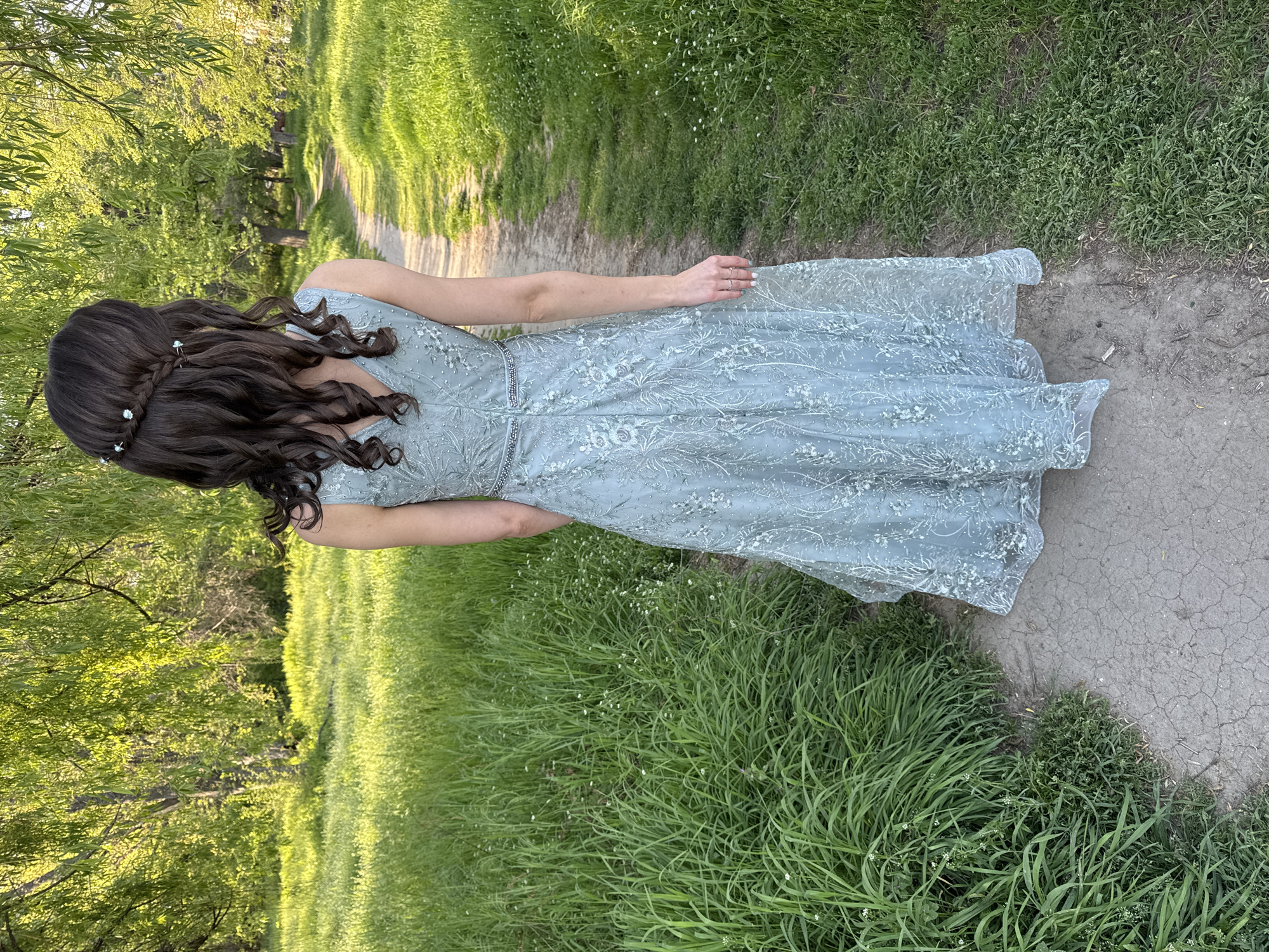 The image depicts a young woman standing on a worn, cracked path bordered by vibrant green grass, possibly in a park or garden. She is wearing a light blue, floor-length dress with intricate lace or floral detailing. Her dark brown hair is styled with a braided section adorned with small, possibly floral, decorations. The path leads into a shadowy area with trees and more greenery, suggesting a trail in a natural setting. The image appears to have been taken outdoors during the day.
Without specific location context within Plovdiv, Bulgaria, I can't pinpoint the exact location.