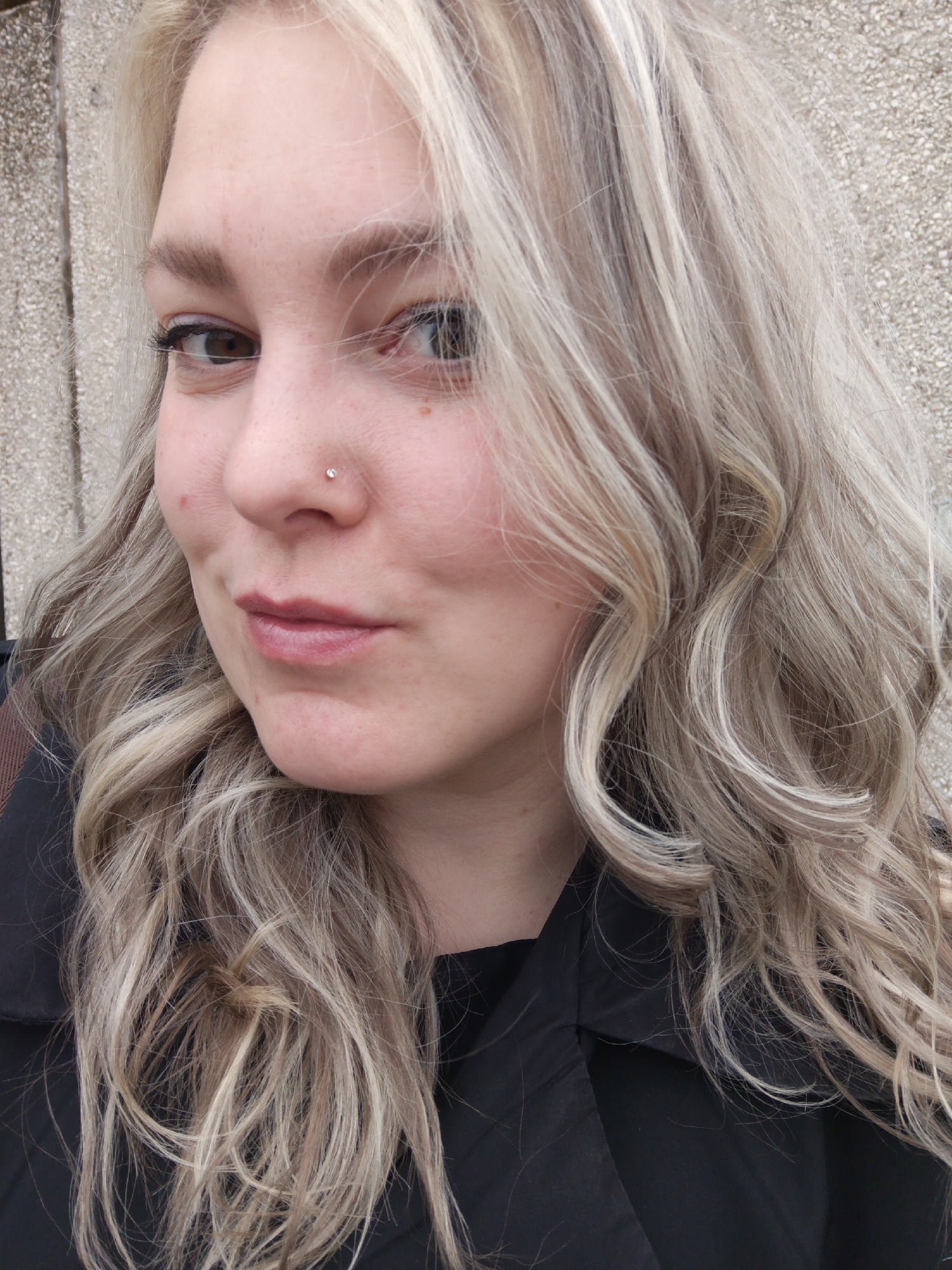 A young woman with blonde hair and brown eyes is facing the camera with a slight smile. She has a nose piercing and several beauty marks on her face. She is wearing a black garment. The background is a neutral concrete wall. The location cannot be confirmed to be in Марковский, Russia.