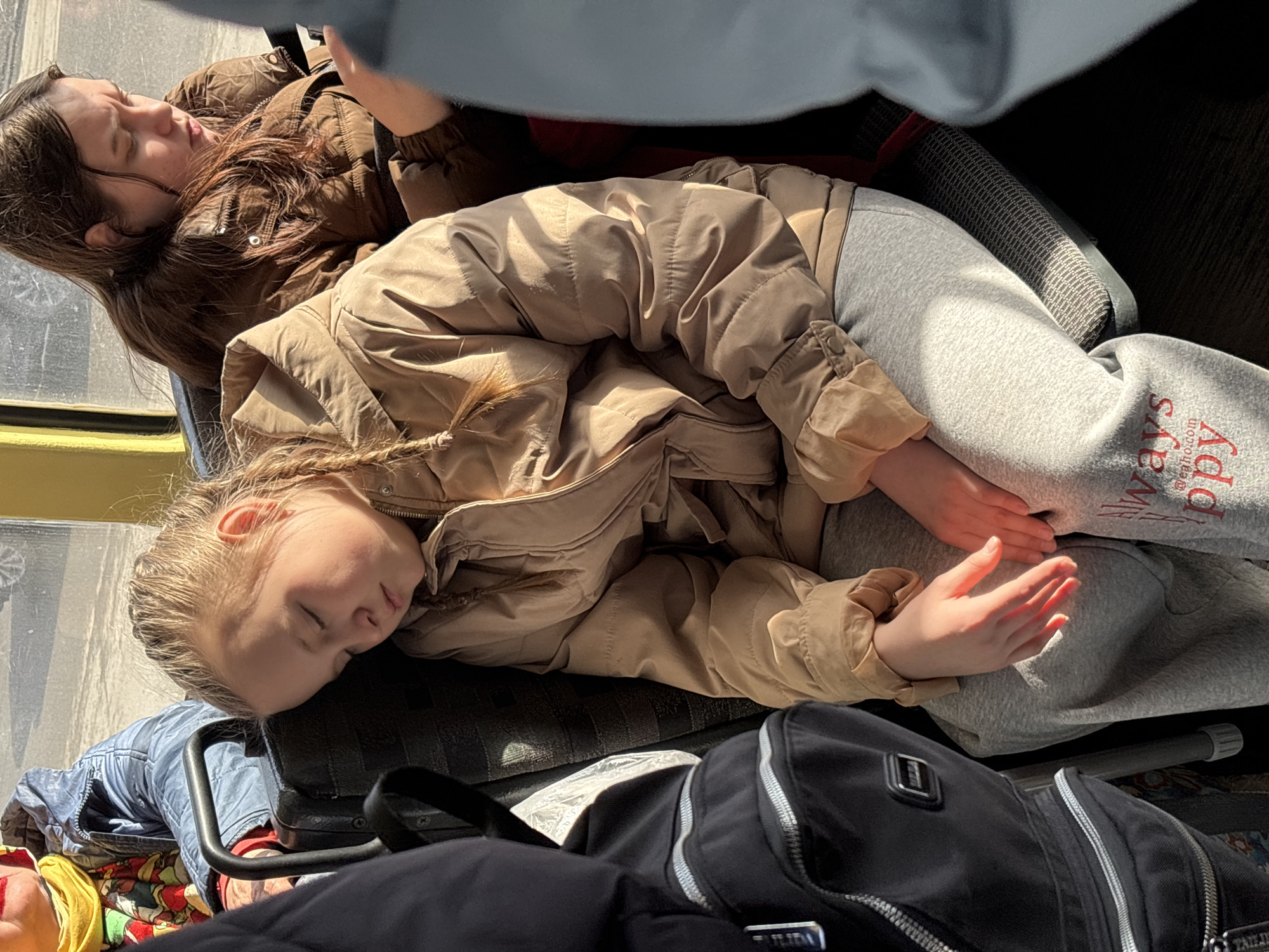 The image depicts two girls sleeping while traveling. The younger girl, with blonde hair in braids, is lying down wearing a beige jacket and grey sweatpants. The sweatpants have some text on them, partially obscured. She is resting her head on a black seat, with her hands placed on her stomach. A black backpack is in the foreground. Behind her, the older girl, with long brown hair, is also asleep. She is wearing a brown puffer jacket. They appear to be seated inside a bus or train, possibly in the city of Izhevsk, Russia, although there are no elements in the image that confirm location. The scene is lit by sunlight.