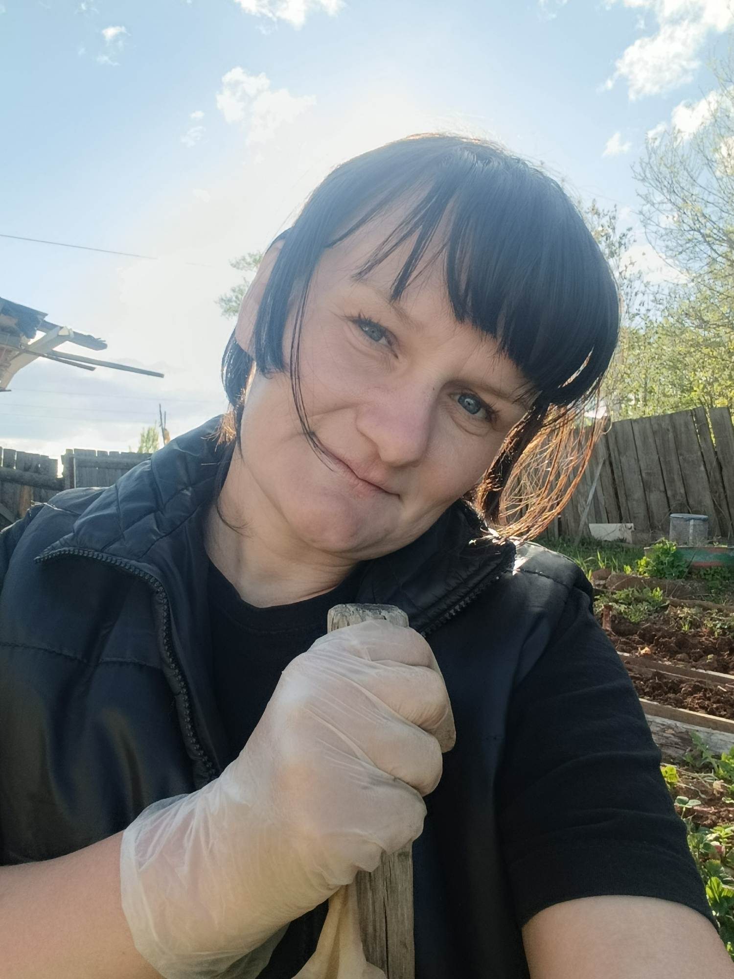 A woman with dark hair and blue eyes is in a garden in Фоки, Russia. She is wearing a black vest and a black shirt. Her right hand is gloved and holding a wooden-handled shovel. The background includes a wooden fence, trees, garden beds, and a blue sky with some clouds. Sunlight illuminates the scene. A structure is partially visible in the top left corner.
