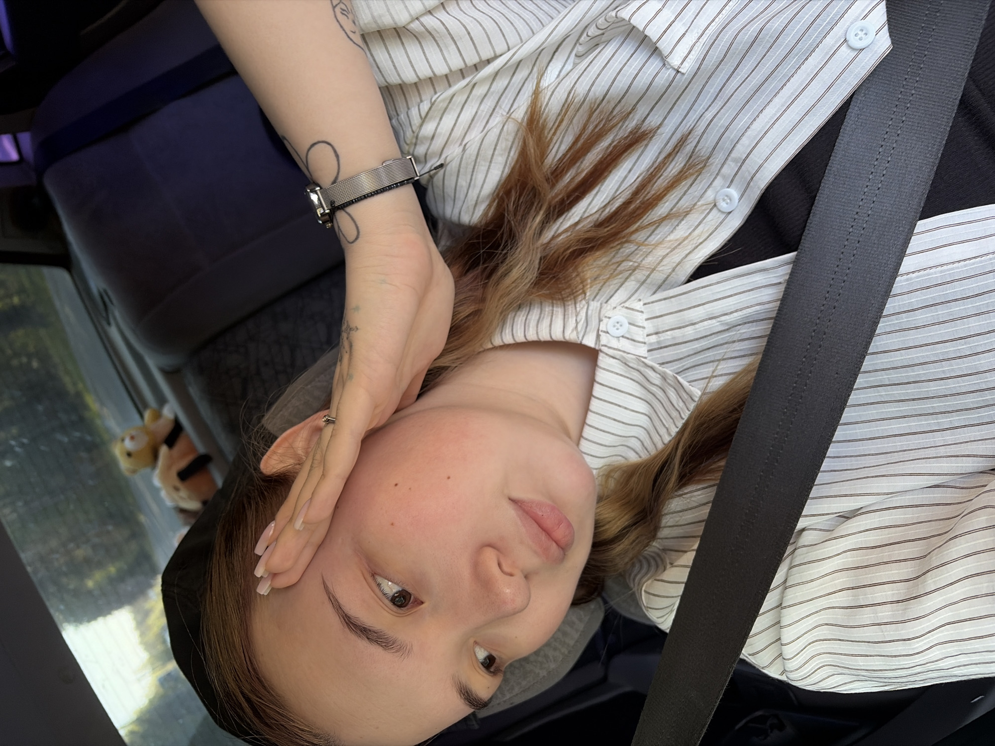 A woman is in a car. She is wearing a striped button-down shirt and a seatbelt. Her arm is raised near her forehead, and she is wearing a watch on that wrist. A stuffed animal is partially visible in the background through the car window. The location is Chaikovsky, Russia.