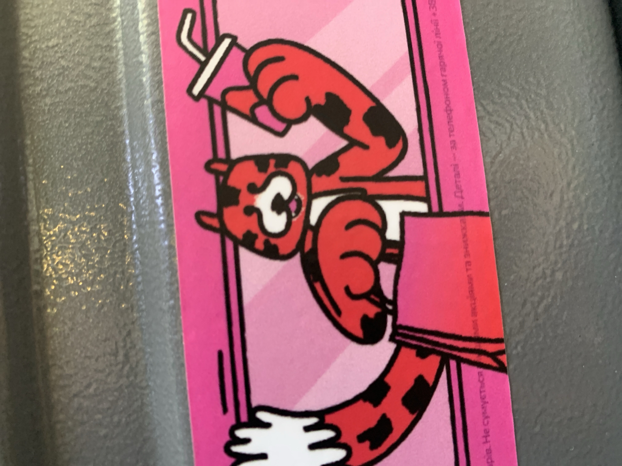 The image is a close-up of a promotional card featuring a cartoon tiger holding a shopping bag and a drink. The tiger is red with black spots and has a white chest and muzzle. The background is pink with diagonal stripes. Text in Ukrainian is visible at the bottom of the card. The card is placed on a textured grey surface. The location of Martynivka, Ukraine is not depicted in the image.