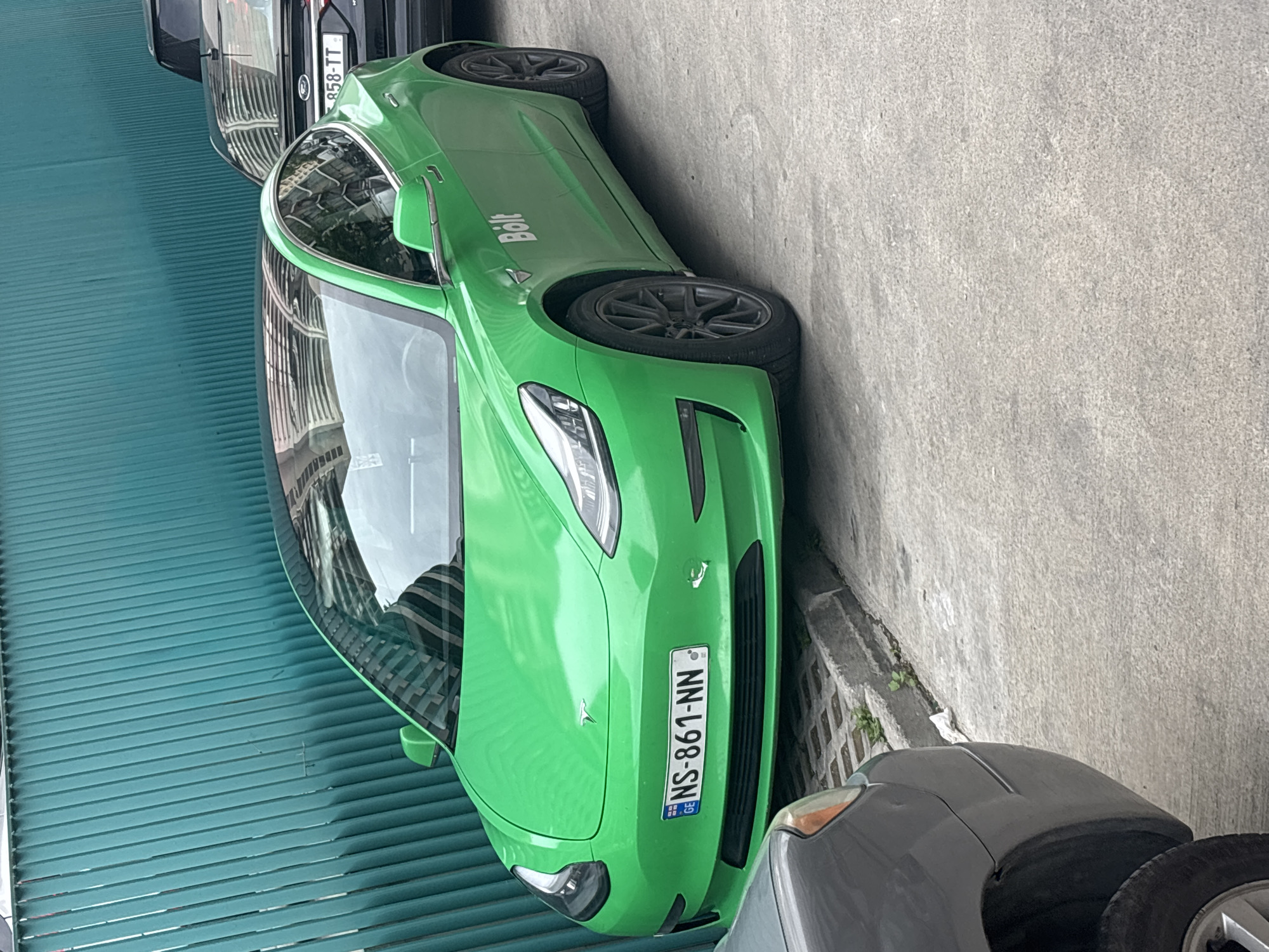 A green Tesla car, with the word "Bolt" on the side, is parked on a city street. The car has a Georgian license plate "NS-861-NN". Another car is partially visible behind the Tesla, and another one in front of the Tesla. The image seems to be taken in the city of Tbilisi, Georgia, given the Georgian license plate on the car. The building on the left has green window slats.